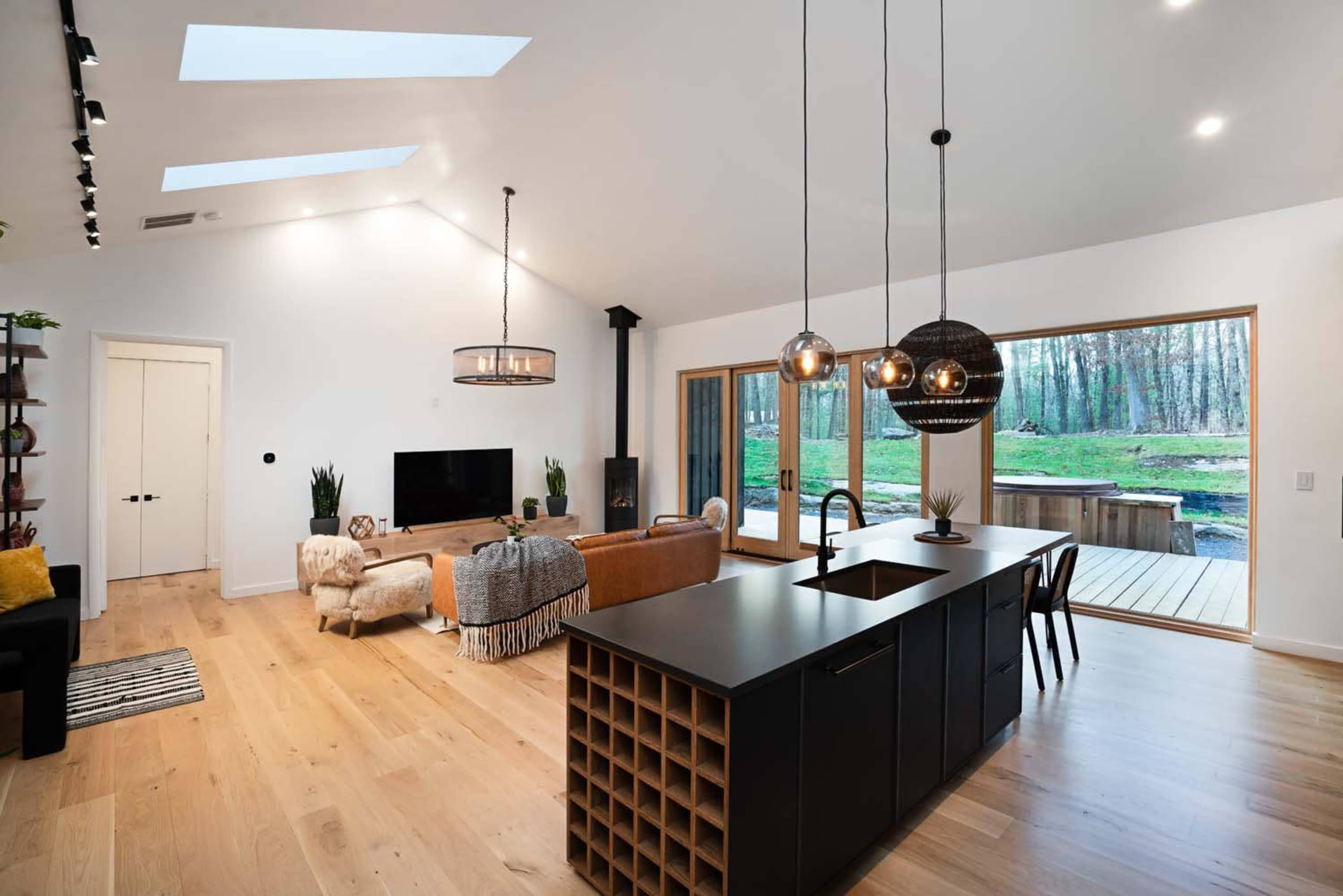 A modern open-concept living space with a kitchen island, a living area, large windows overlooking a wooded area, and a doorway leading outside.