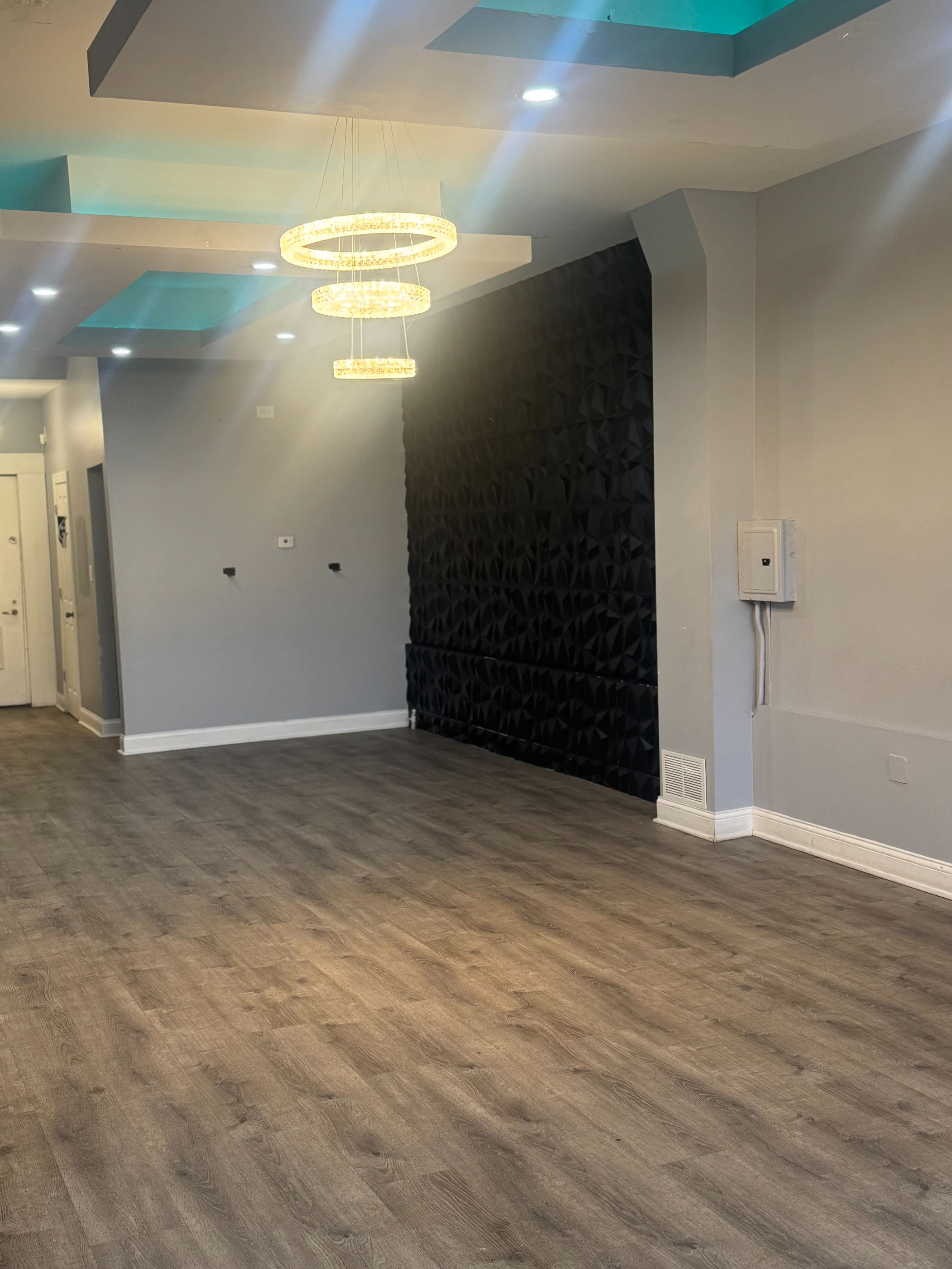 The image shows a modern interior space with a wooden floor, a textured black feature wall, and a stylish chandelier hanging from the ceiling.
