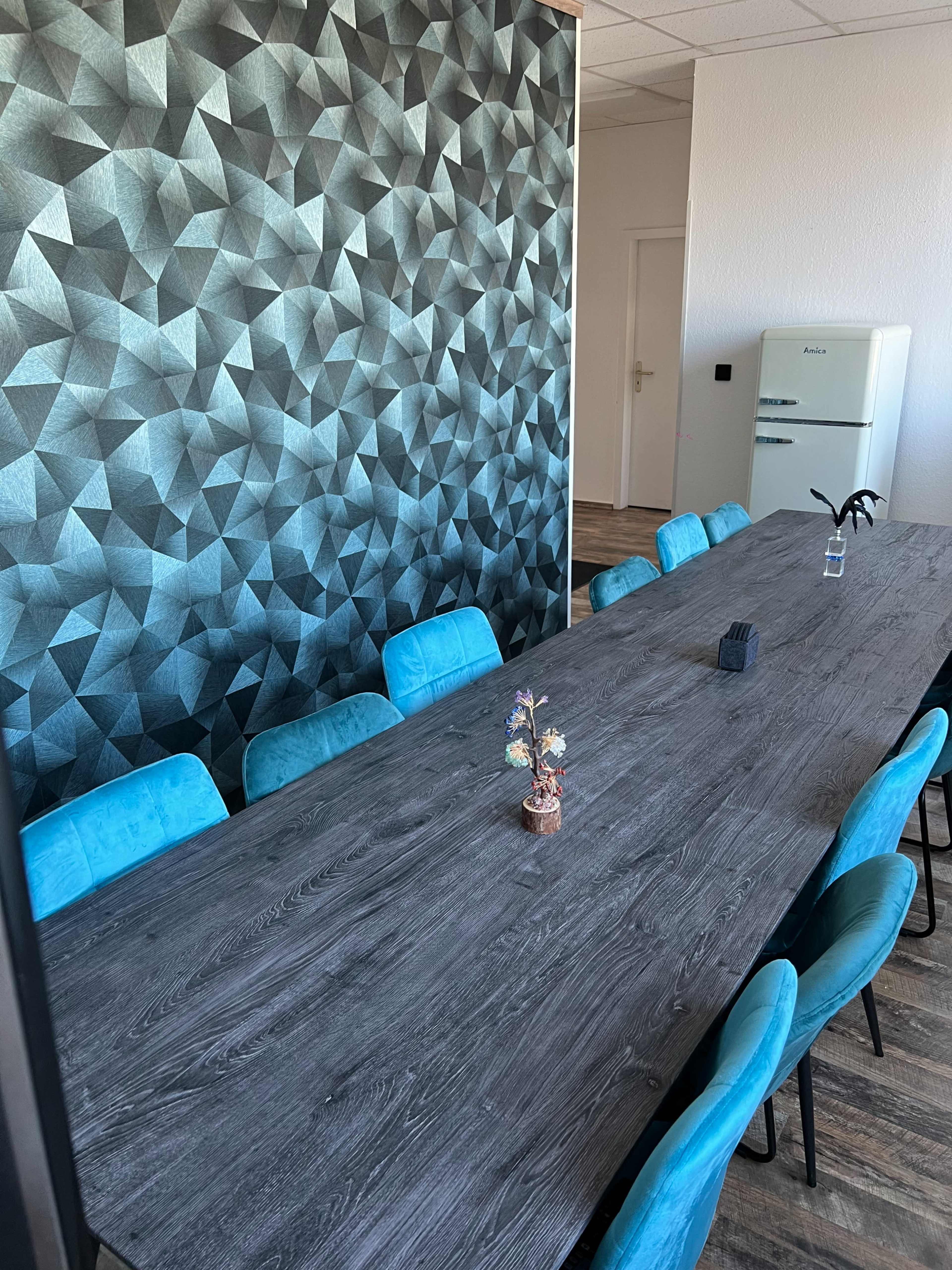 A long wooden conference table is surrounded by blue chairs, with a geometric-patterned wall and a small decorative plant on the table.