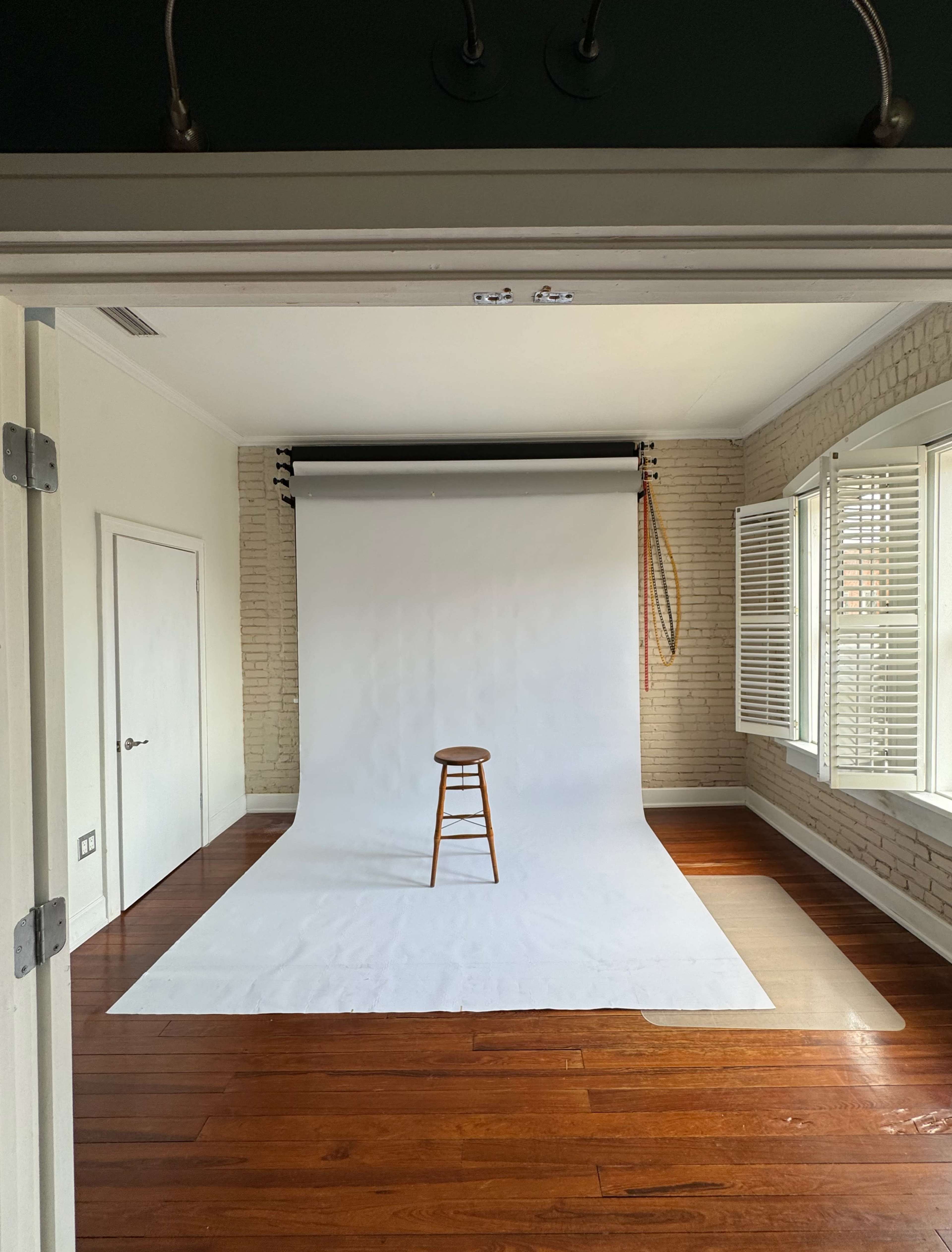 A photography studio with a wooden stool centered on a white backdrop and natural light streaming through window blinds.