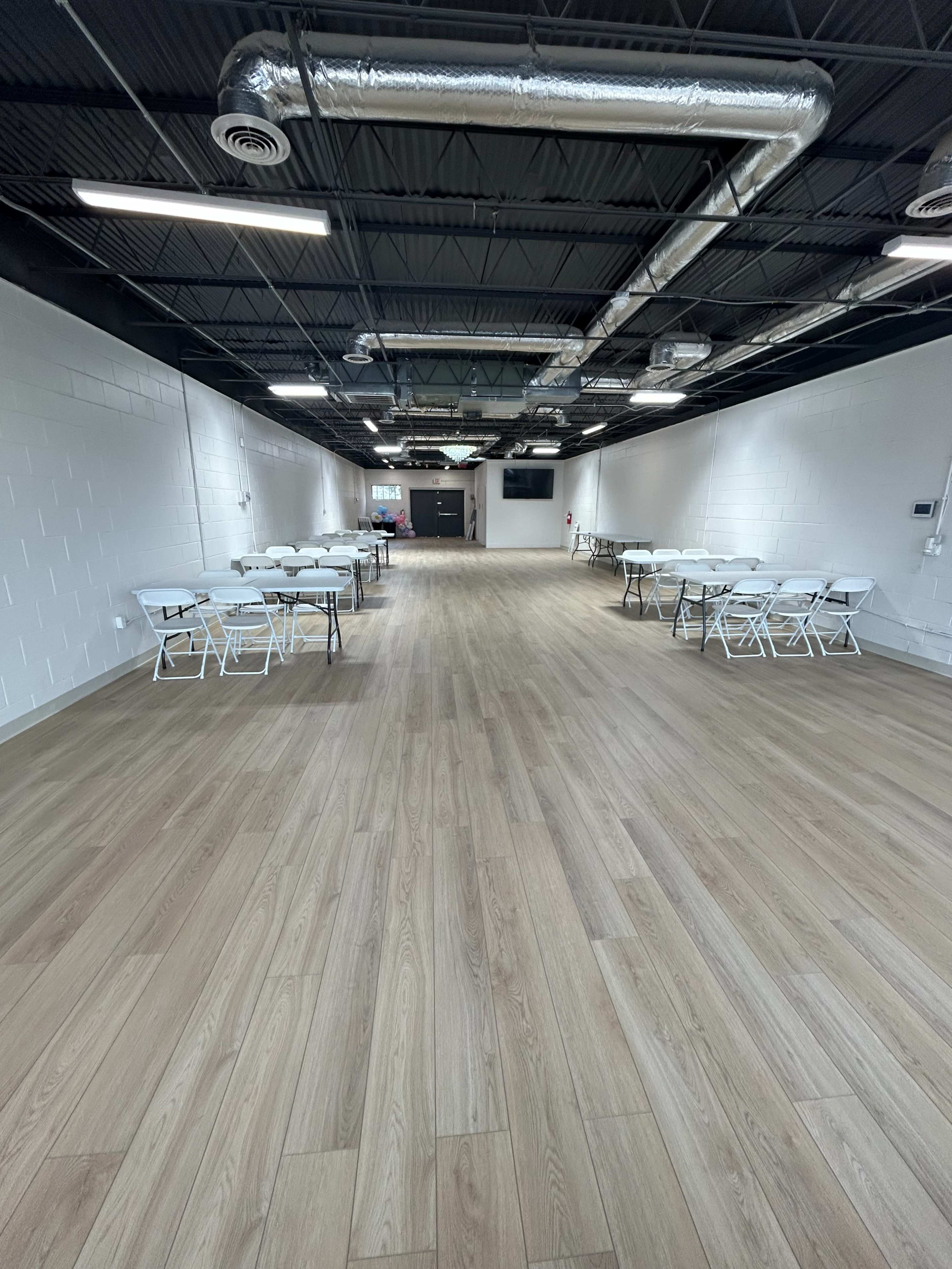 The image shows a spacious, empty room featuring several rows of white folding tables and chairs arranged neatly on a wooden floor.