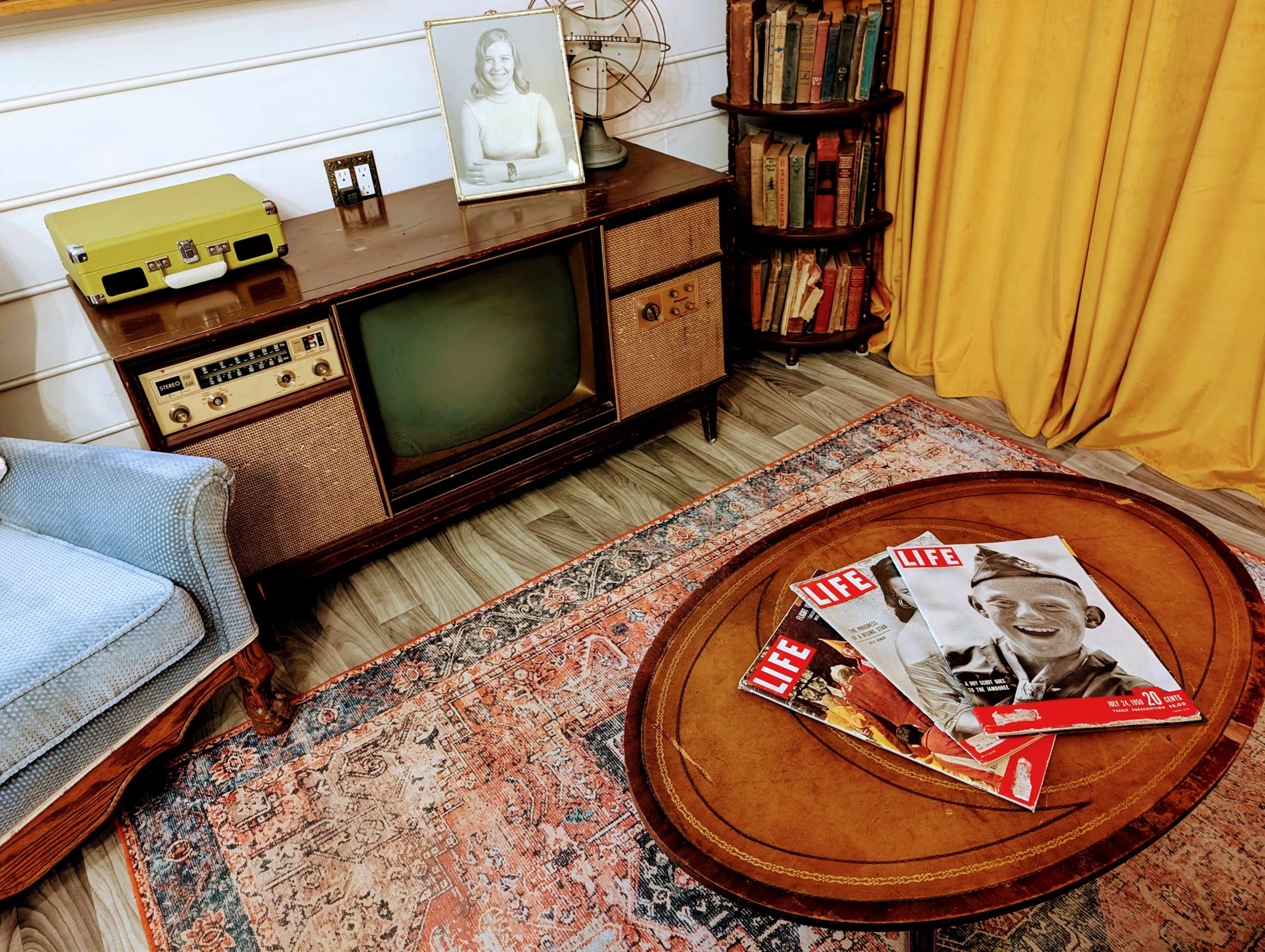 The scene features a vintage living room setup with a retro television, an old photograph in a frame, a wooden coffee table displaying LIFE magazines, and a bookshelf in the background.