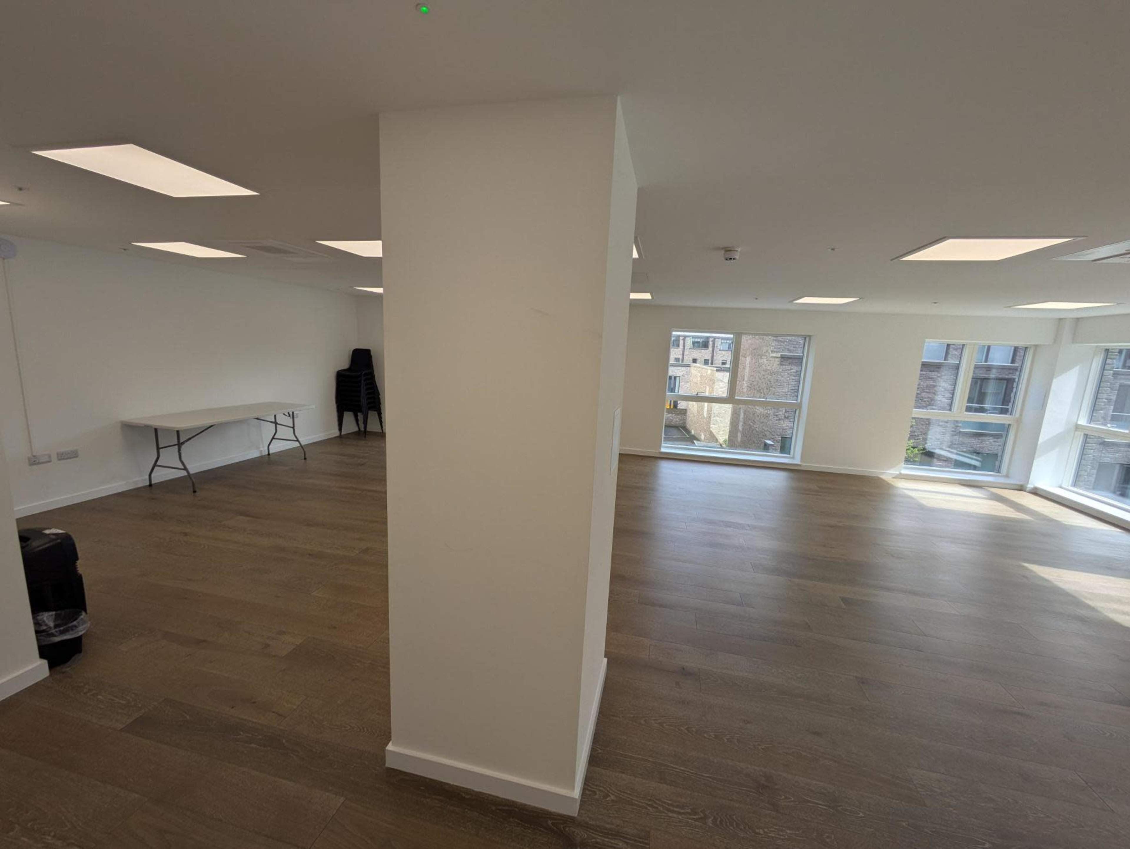 The image shows a spacious, well-lit room with large windows, wooden flooring, and a partitioning column in the center.