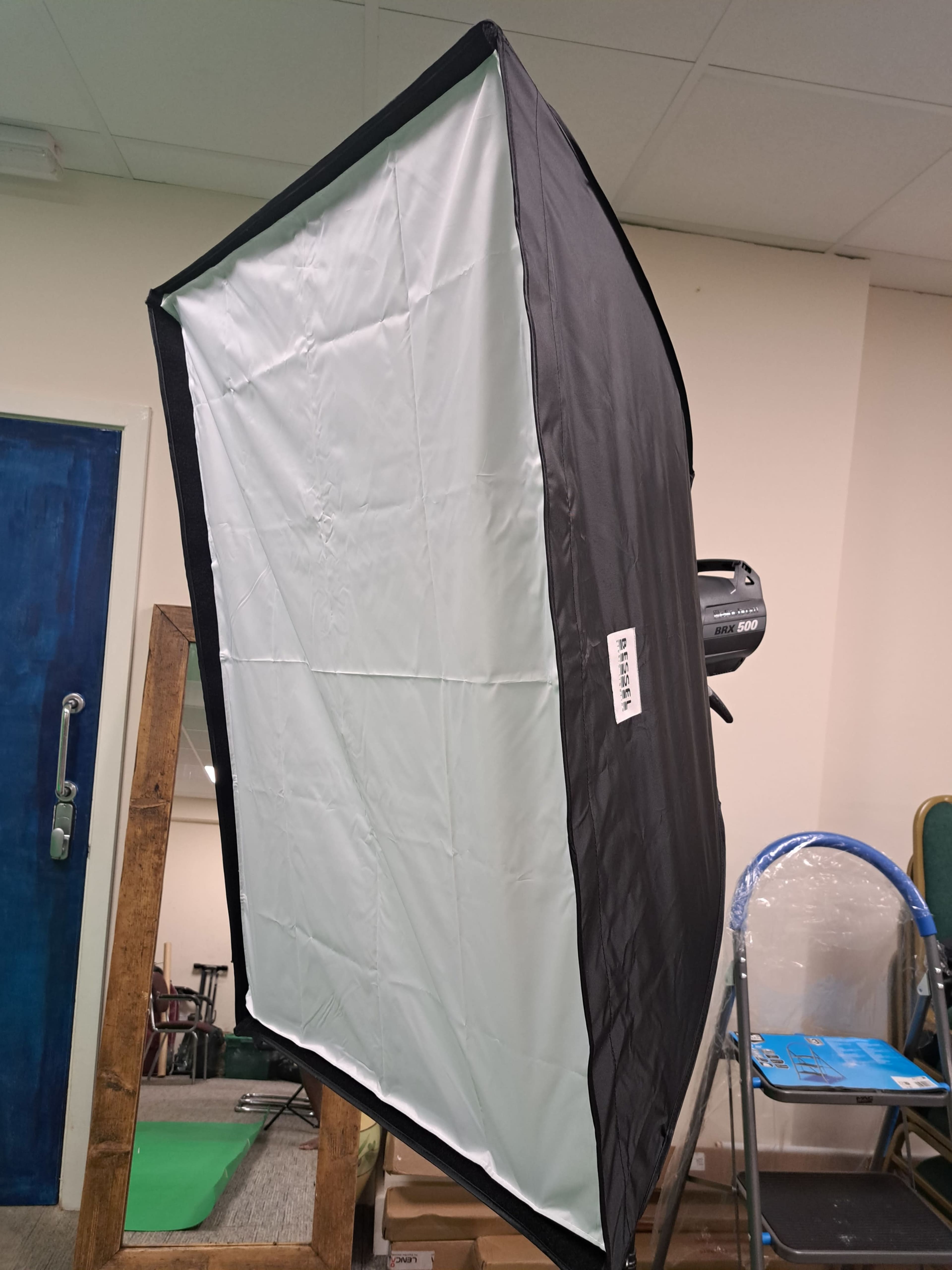 A rectangular softbox with a white interior and a black exterior, positioned in a room with a blue door and a wooden mirror frame.