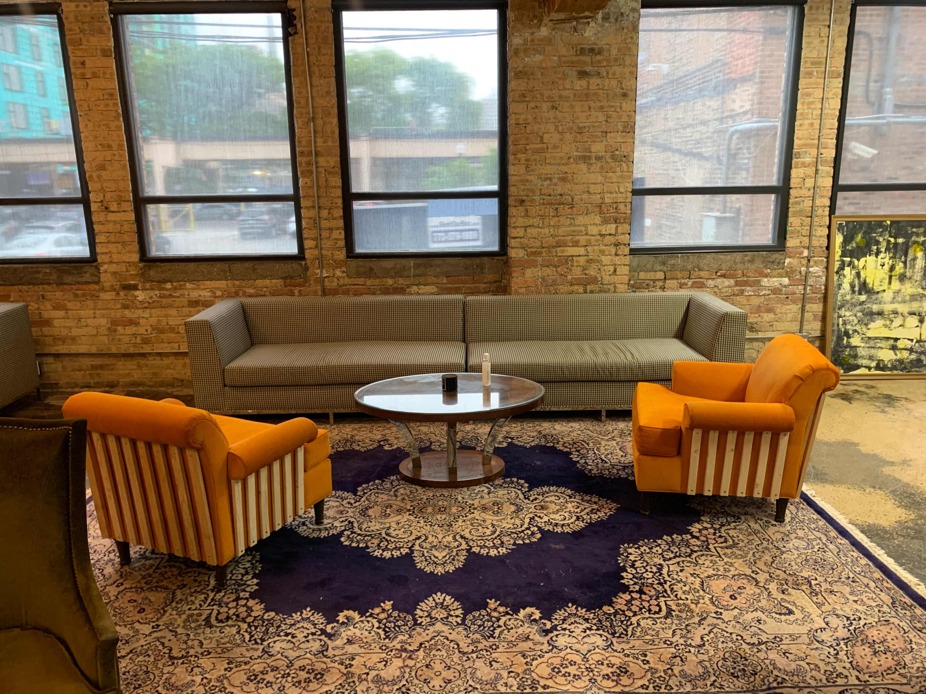 A living area with a light gray sofa, two orange chairs, and a round glass coffee table on a patterned rug, situated in a room with large windows.