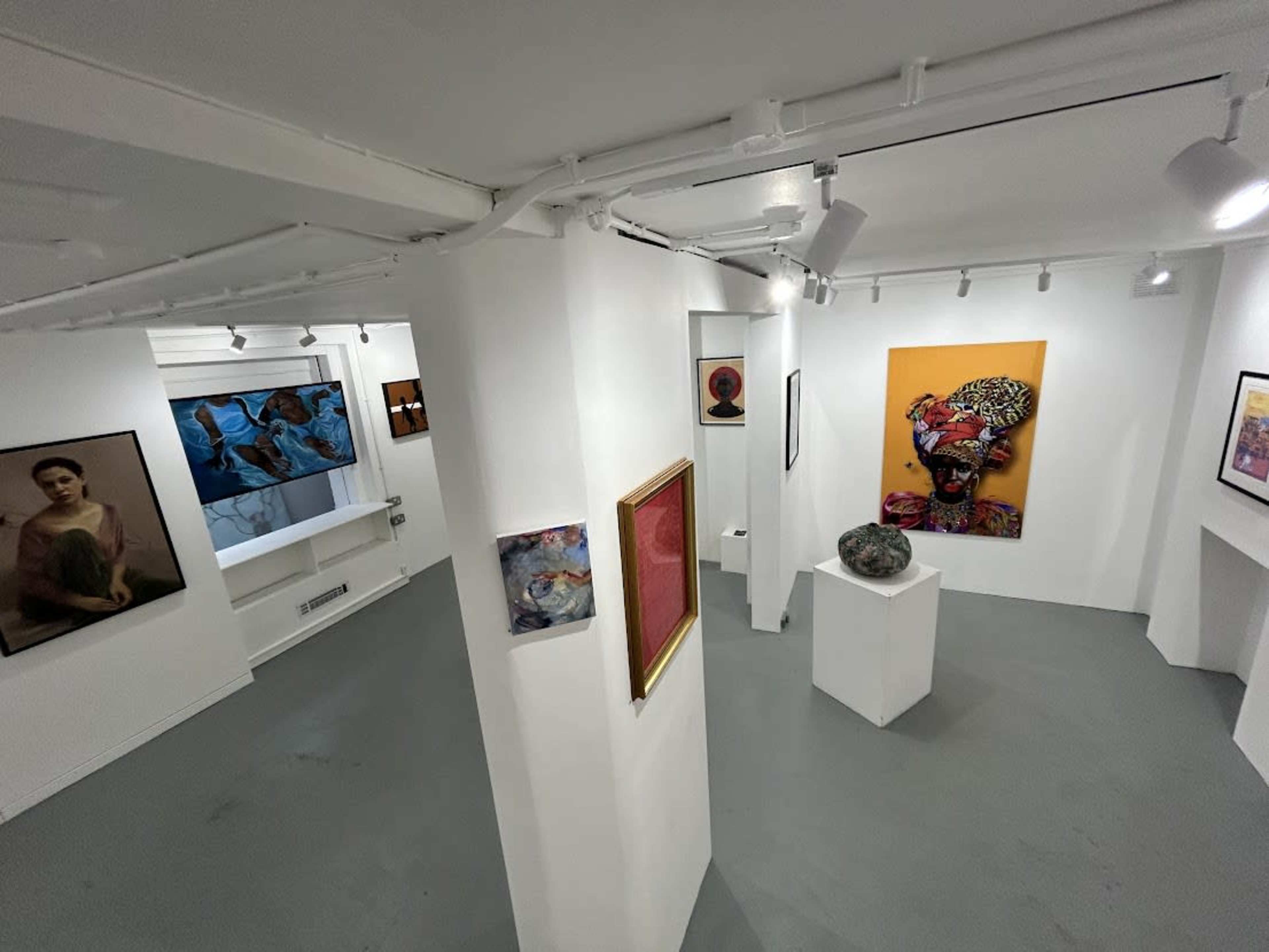 An interior view of an art gallery with white walls displaying various artworks, including a colorful portrait, a sculpture on a pedestal, and framed pieces.