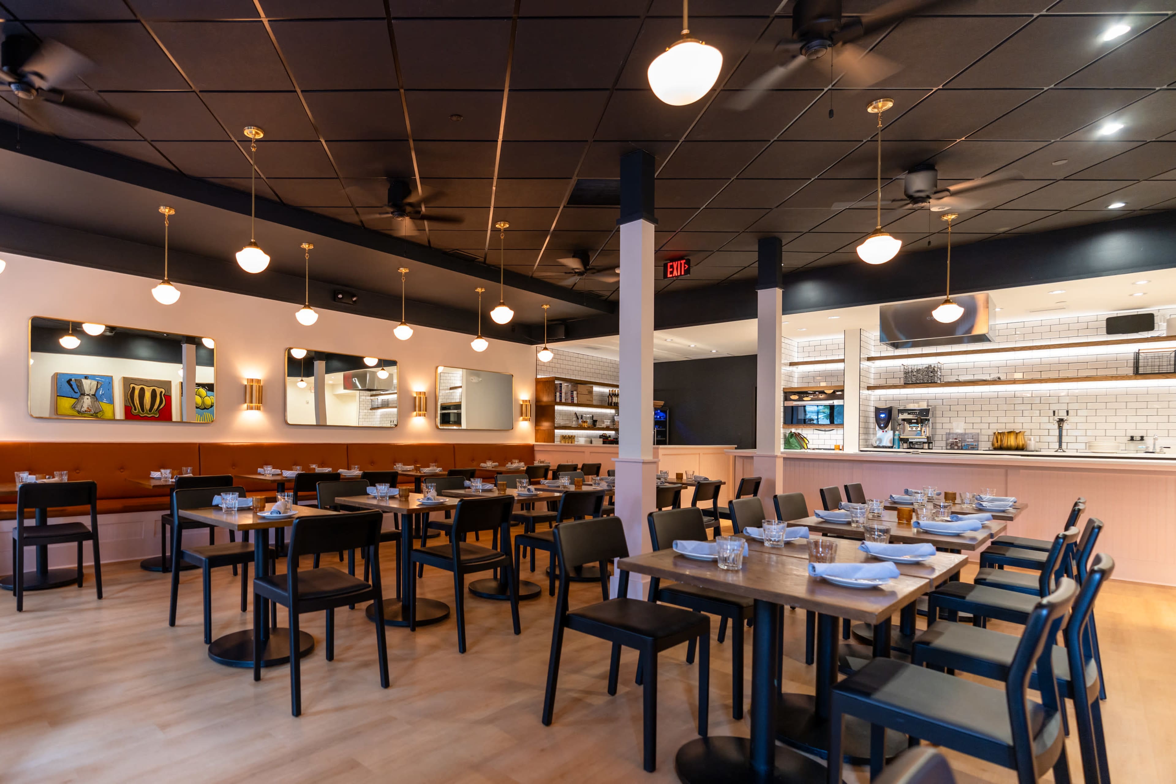The image shows a modern restaurant interior with tables set for dining, soft lighting from pendant lamps, and a visible open kitchen area in the background.