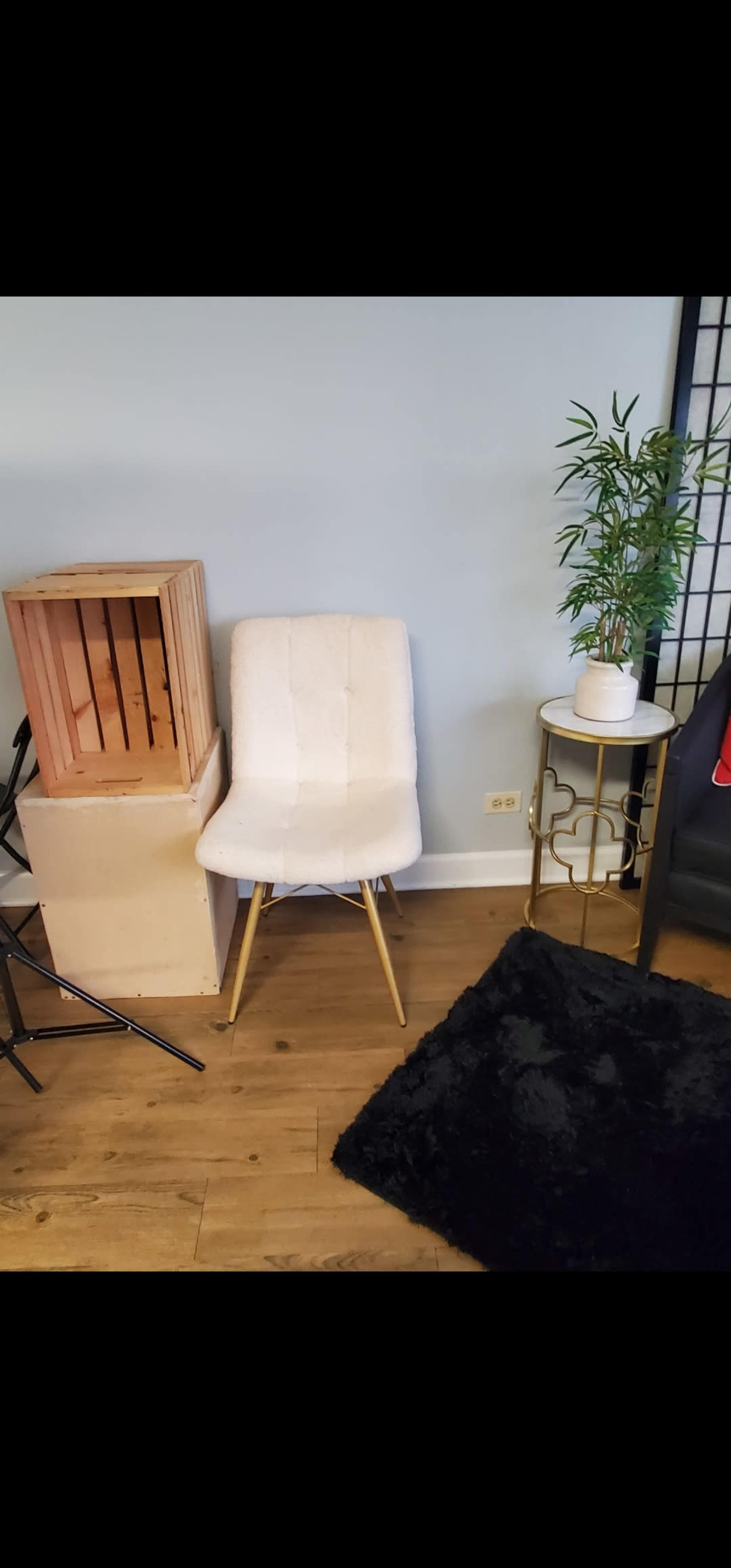 A white upholstered chair next to a wooden crate, a small round table with a plant on it, and a black rug on a wooden floor.