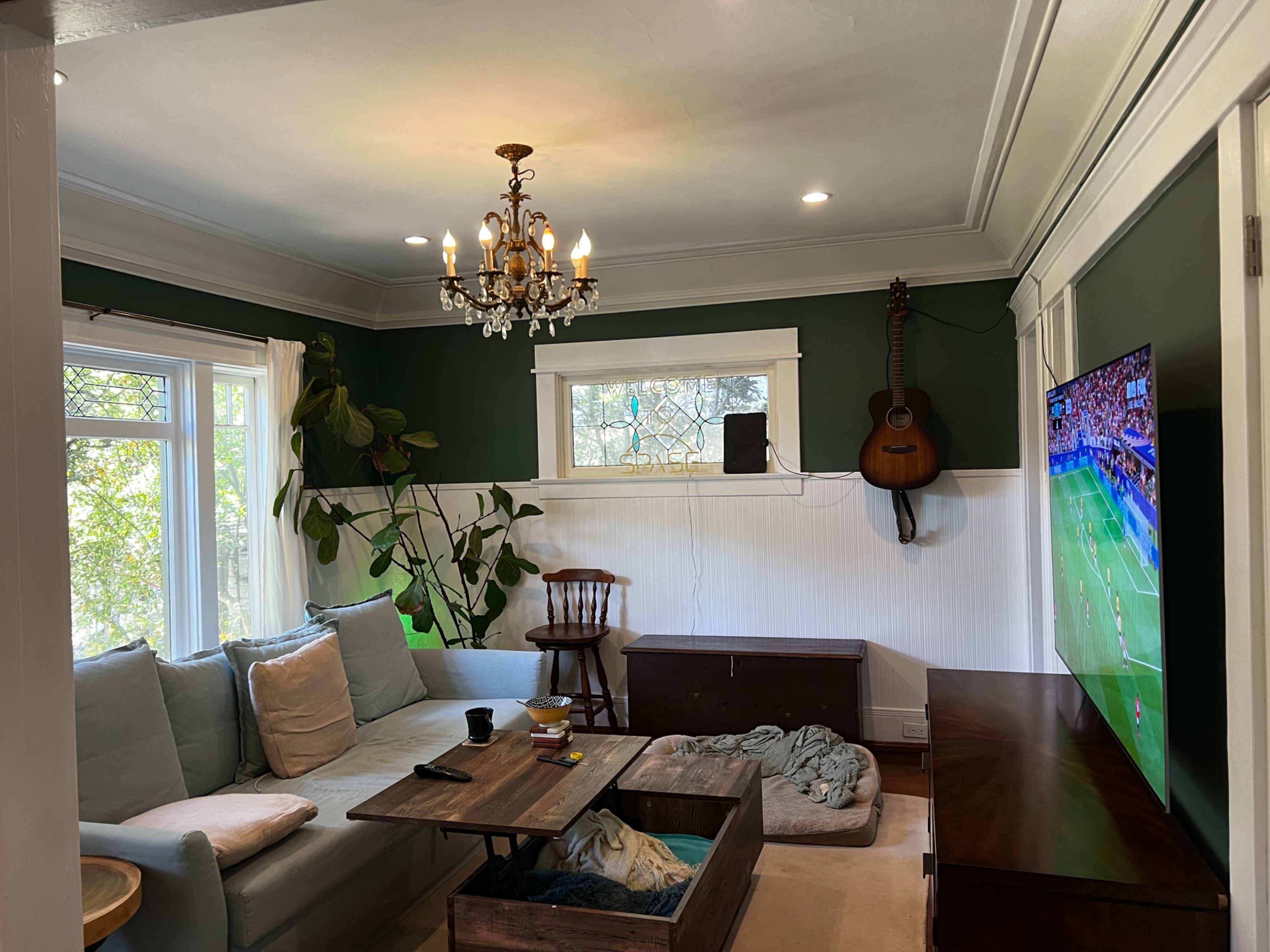 The image shows a living room with a sectional sofa, a wooden coffee table, a guitar hanging on the wall, and a television displaying a sports game.