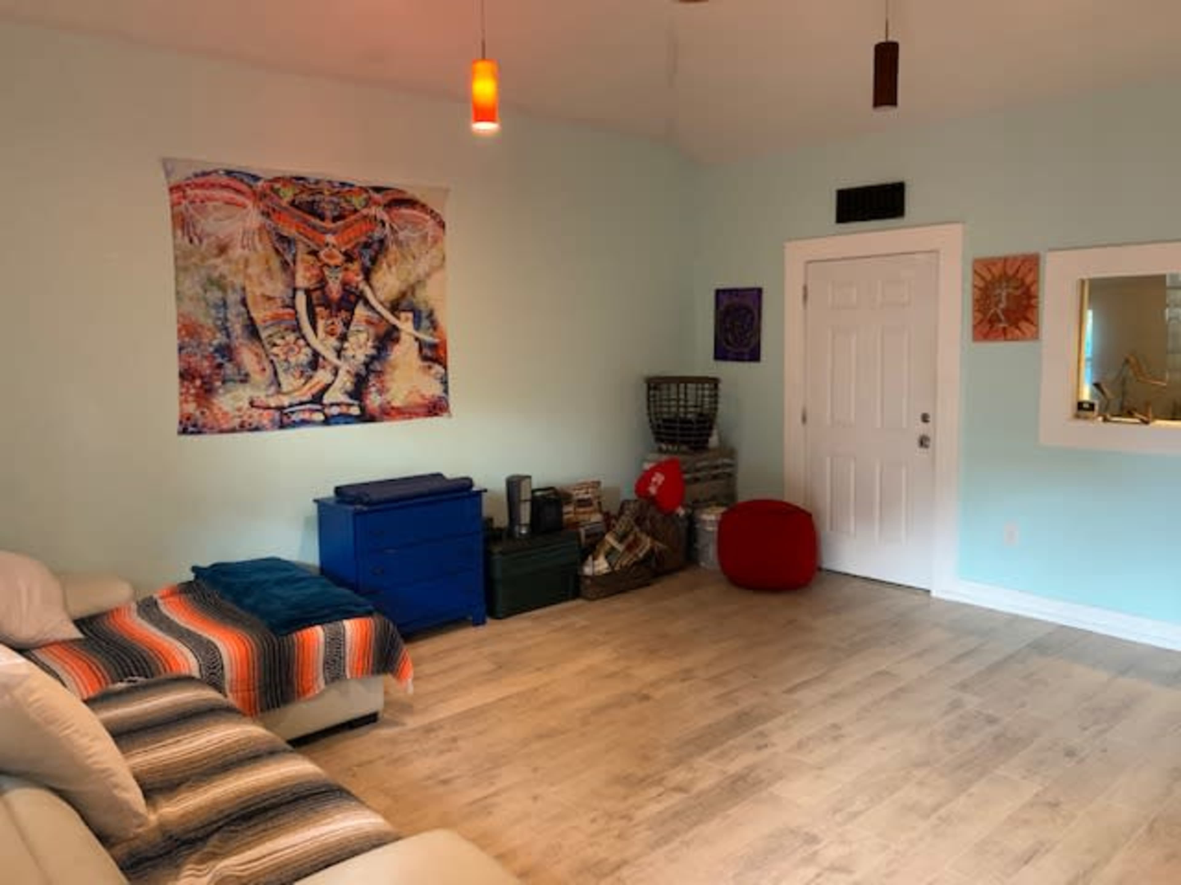 The room features a light blue wall with an elephant tapestry, a white couch with colorful throw pillows, and a door leading outside.