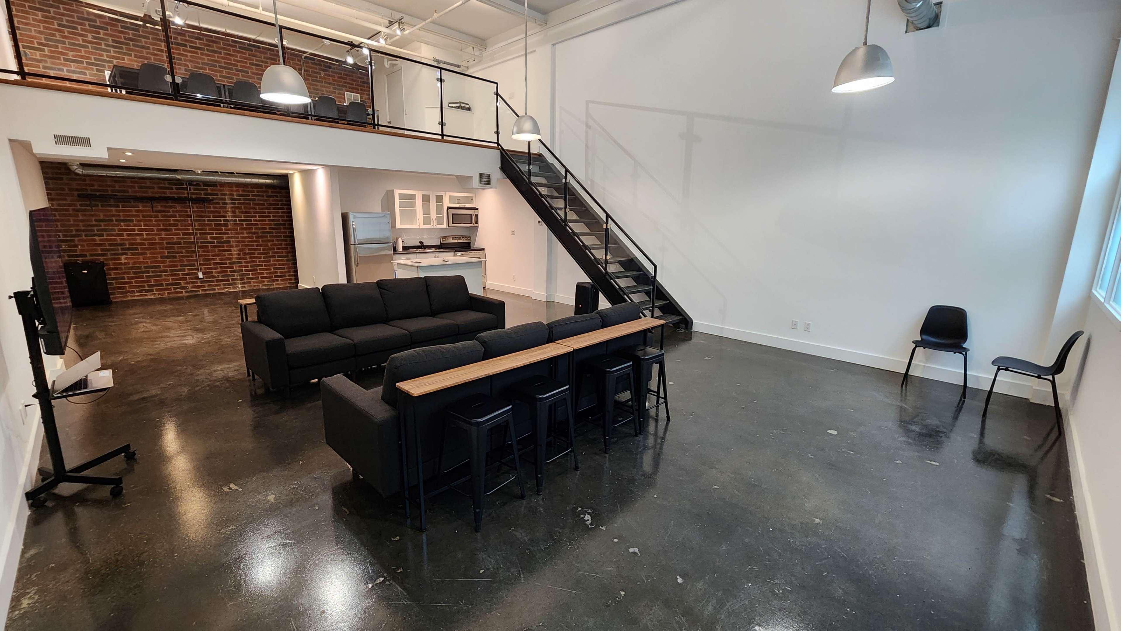 A spacious room featuring a black sectional sofa, a dining table with black chairs, a kitchen area in the background, and a staircase leading to a second level.