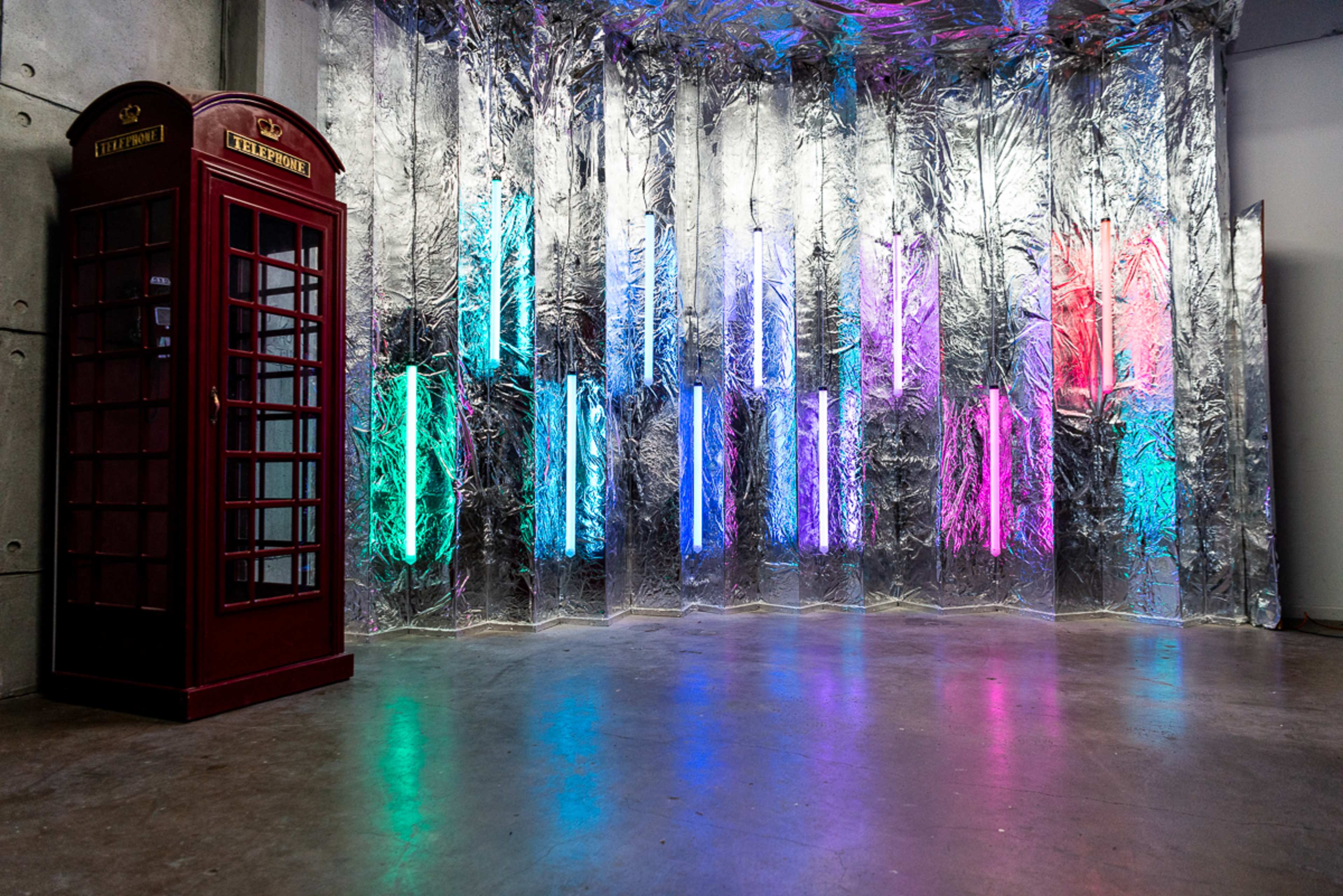 A red telephone booth stands next to a wall covered in shiny foil with vertical neon lights in various colors.