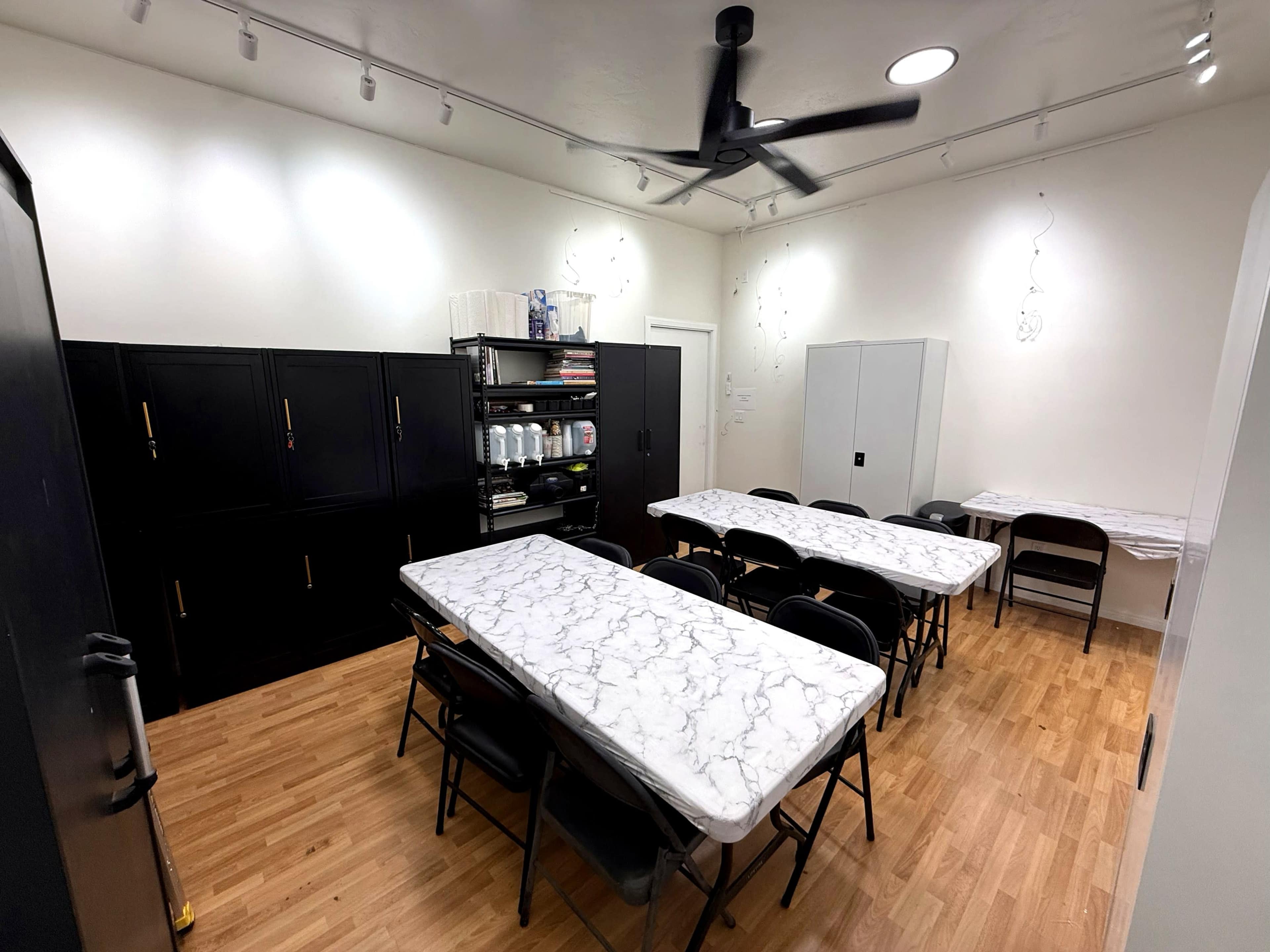 The image shows a classroom or meeting space with multiple tables arranged in a U-shape, surrounded by black folding chairs, and equipped with shelving units on one side.