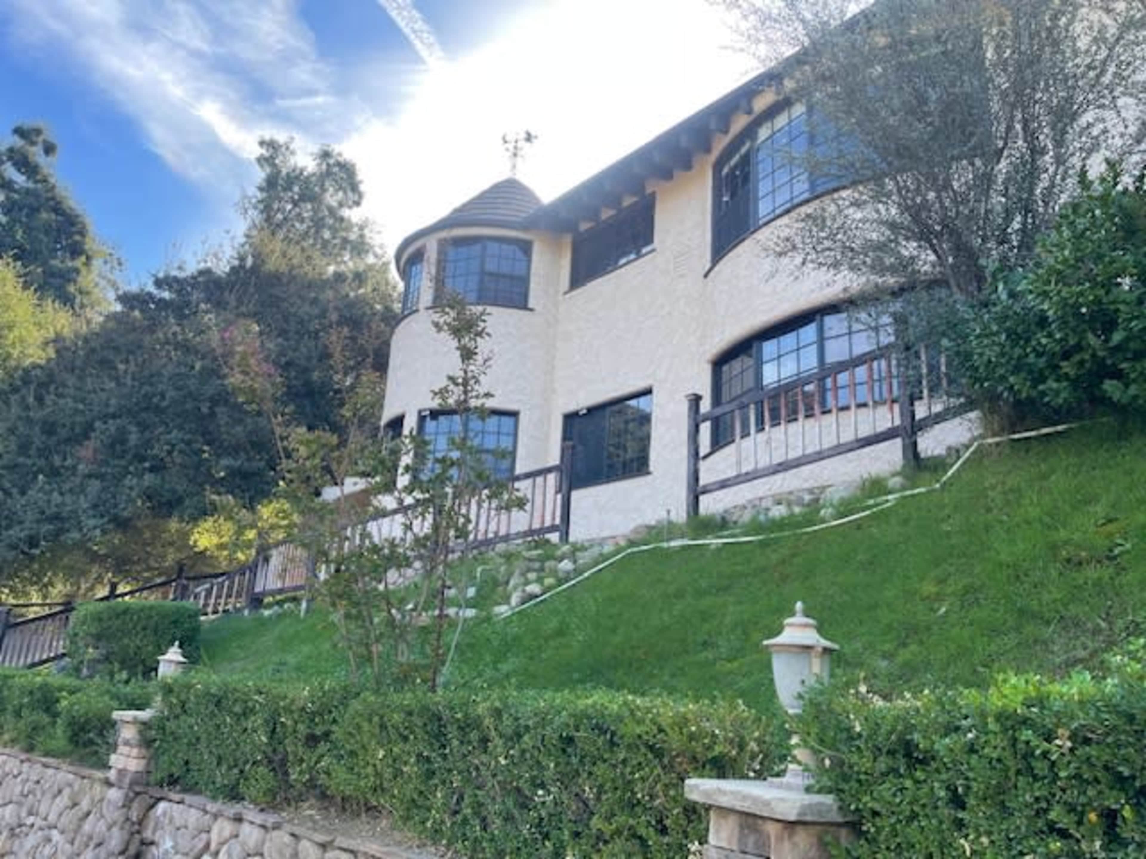 A multi-layered house with large windows and a stone wall is situated on a hillside surrounded by greenery.
