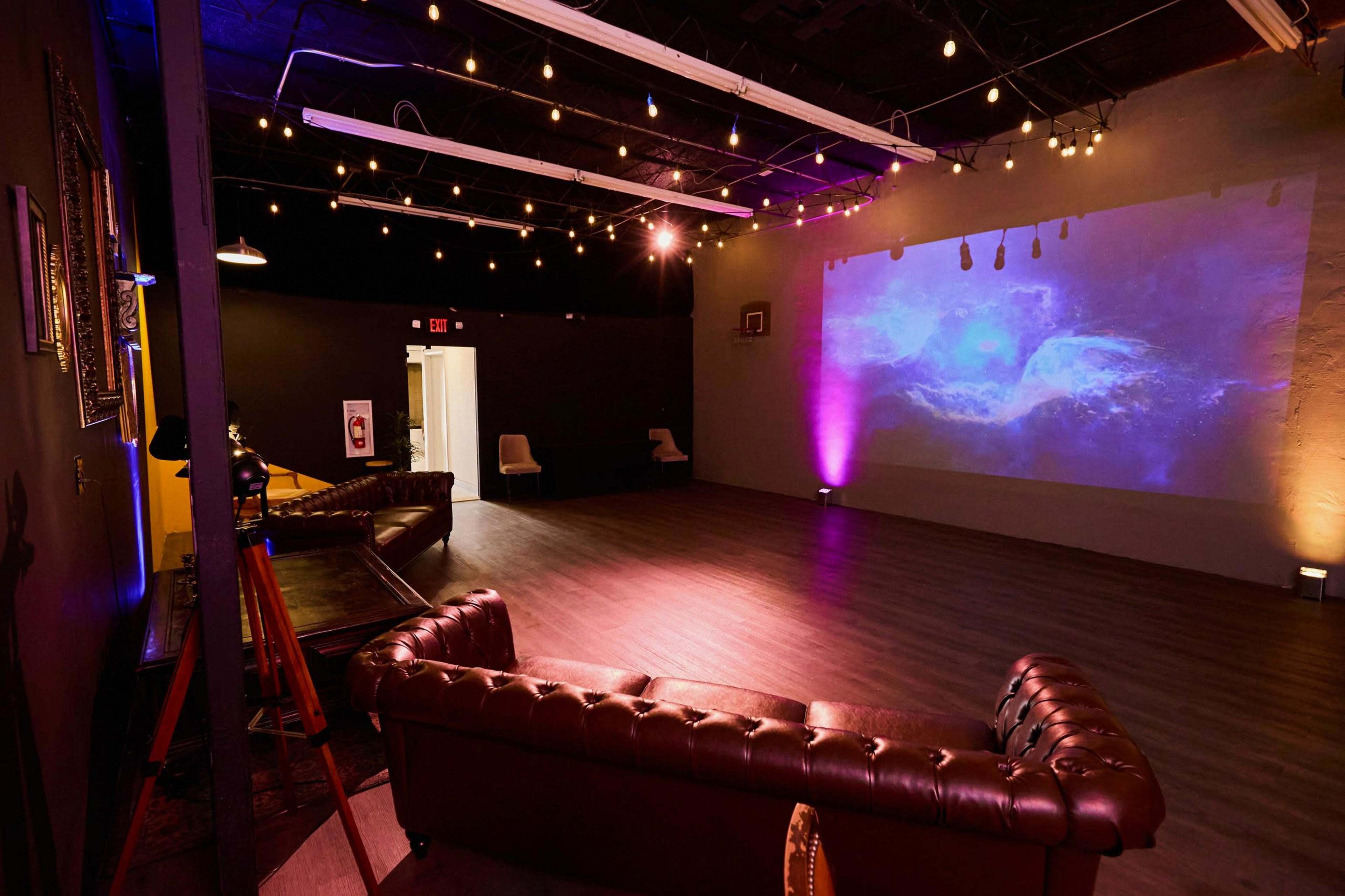 The image shows a spacious, dimly lit room with a projection on the wall, a leather couch, and string lights overhead.