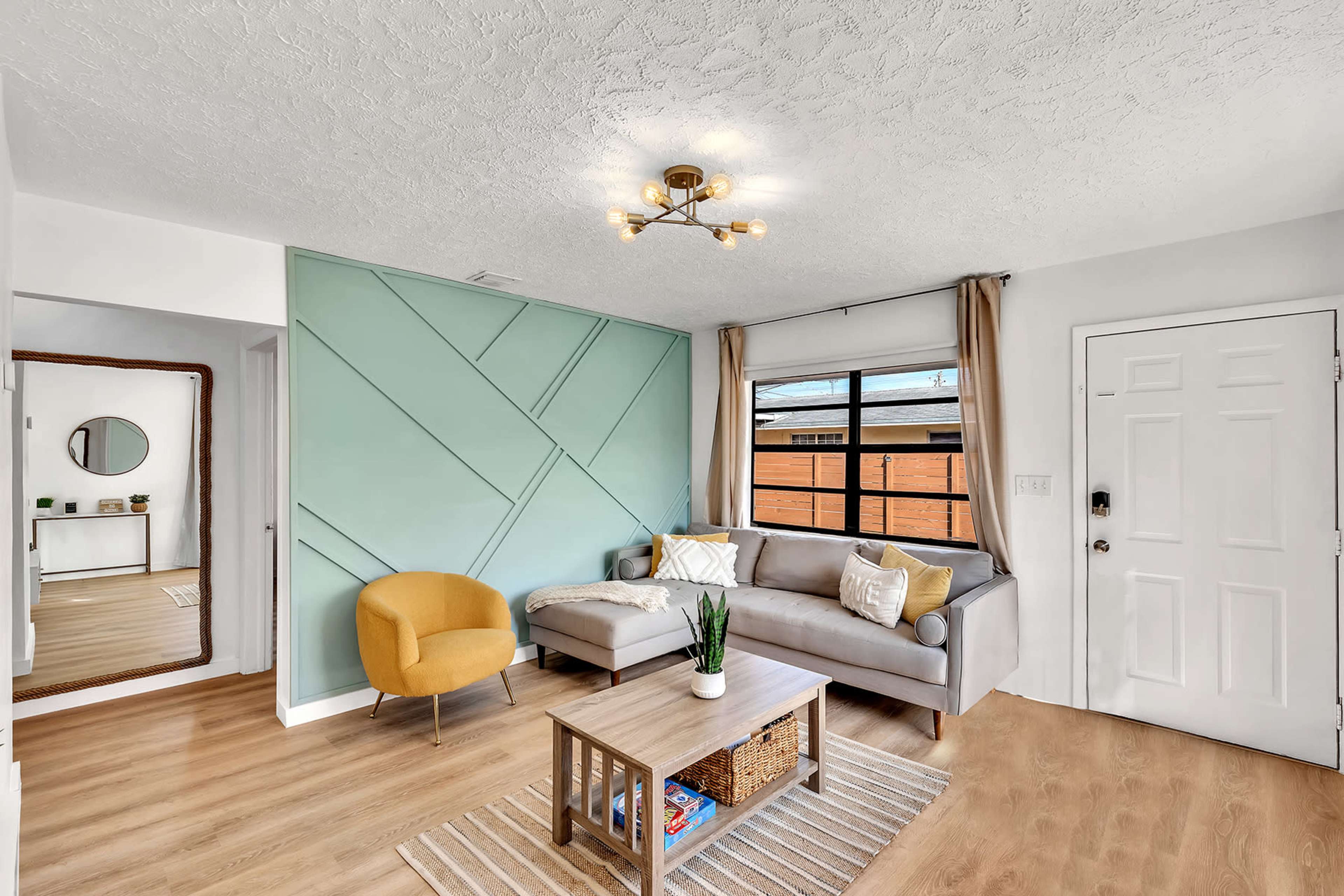 A modern living room with a gray sectional sofa, a yellow accent chair, a wooden coffee table with storage, and light-colored walls featuring a geometric design.