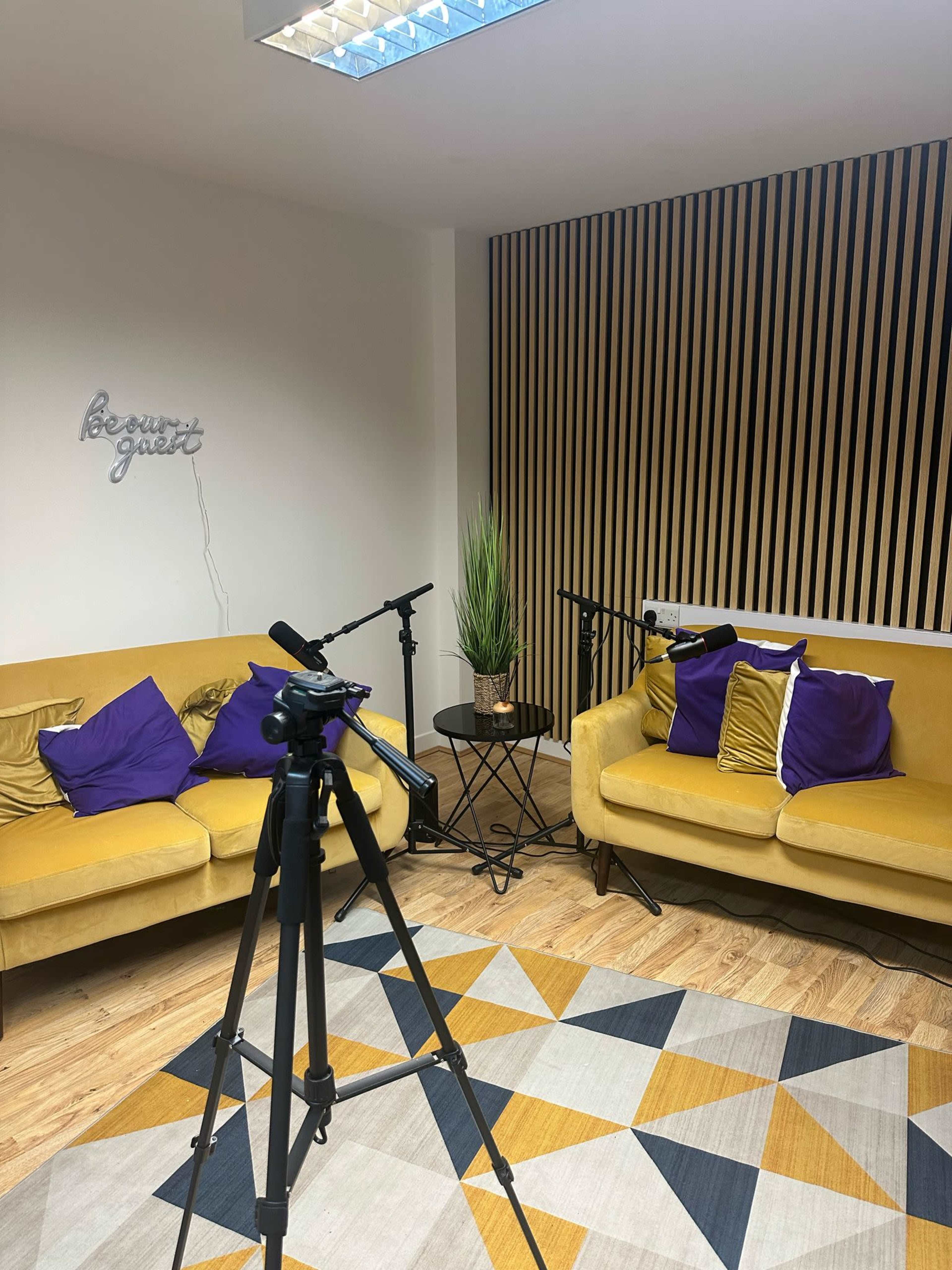 The image shows a small recording studio with two yellow sofas, purple cushions, a round table, and a microphone setup on a tripod.