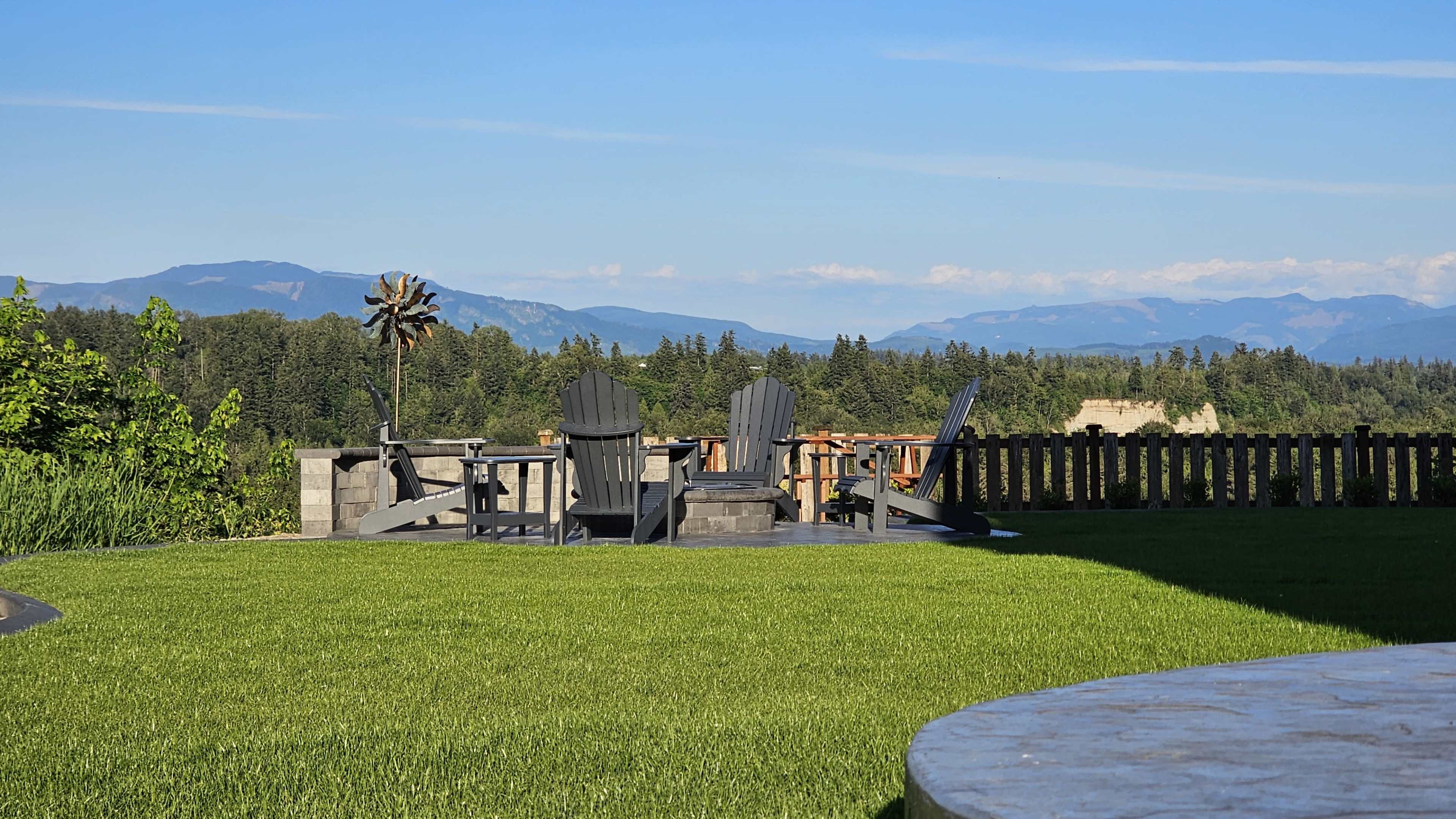 A clear view of a lawn with black Adirondack chairs and a wind spire, set against a backdrop of mountains and a blue sky.