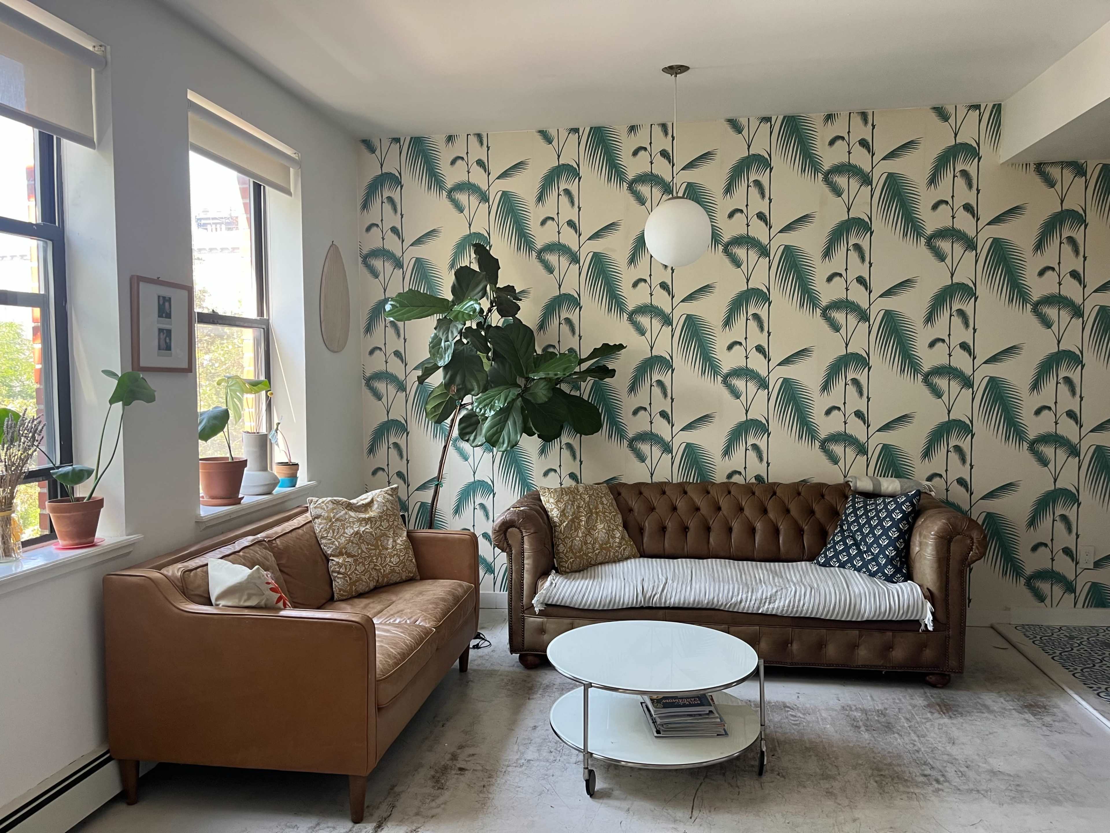 The image shows a living room with two sofas, one brown leather and the other a lighter fabric, surrounded by green plants and a patterned wallpaper featuring palm leaves.