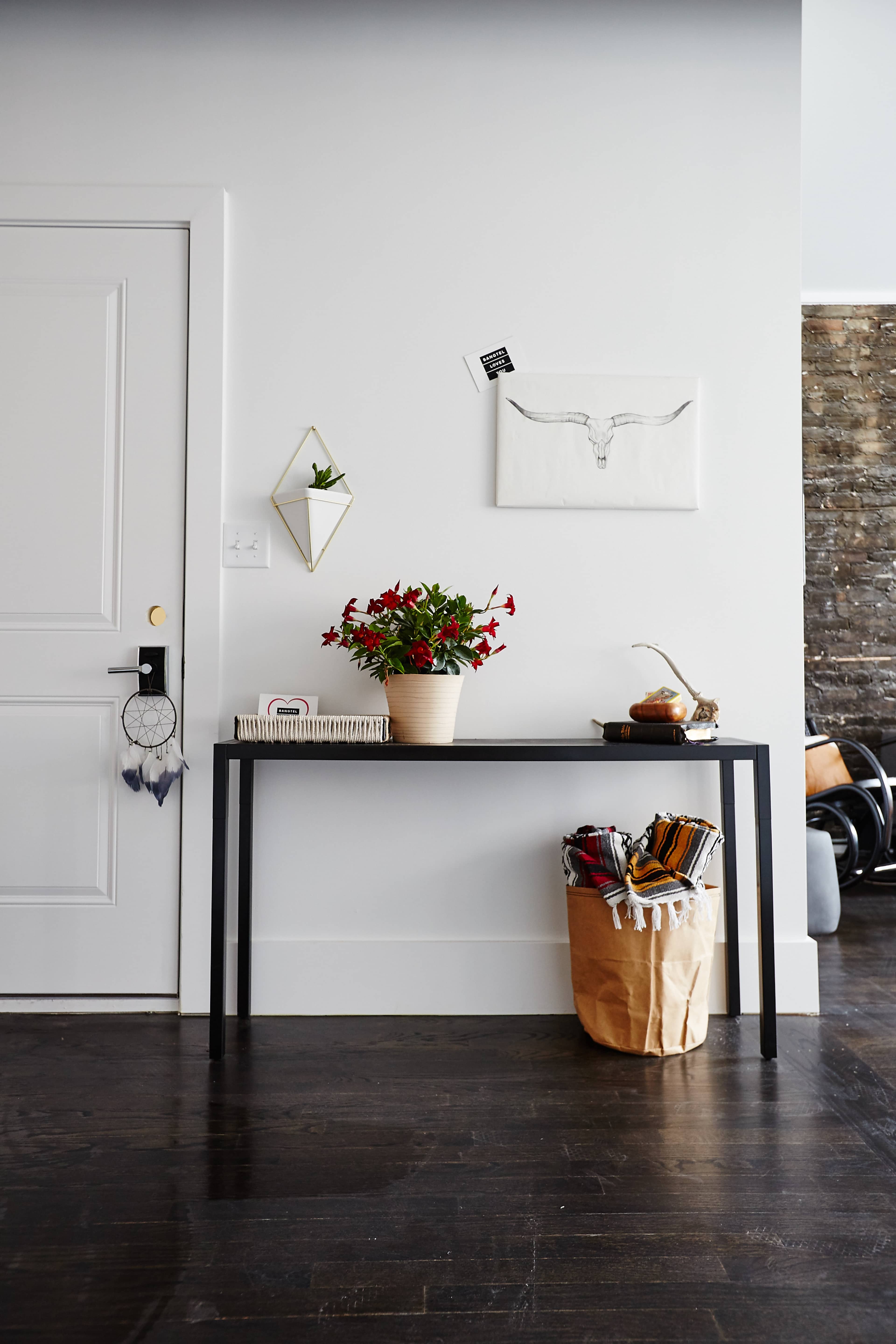 A black console table is positioned against a white wall, adorned with a potted plant, decorative items, and a basket filled with blankets.