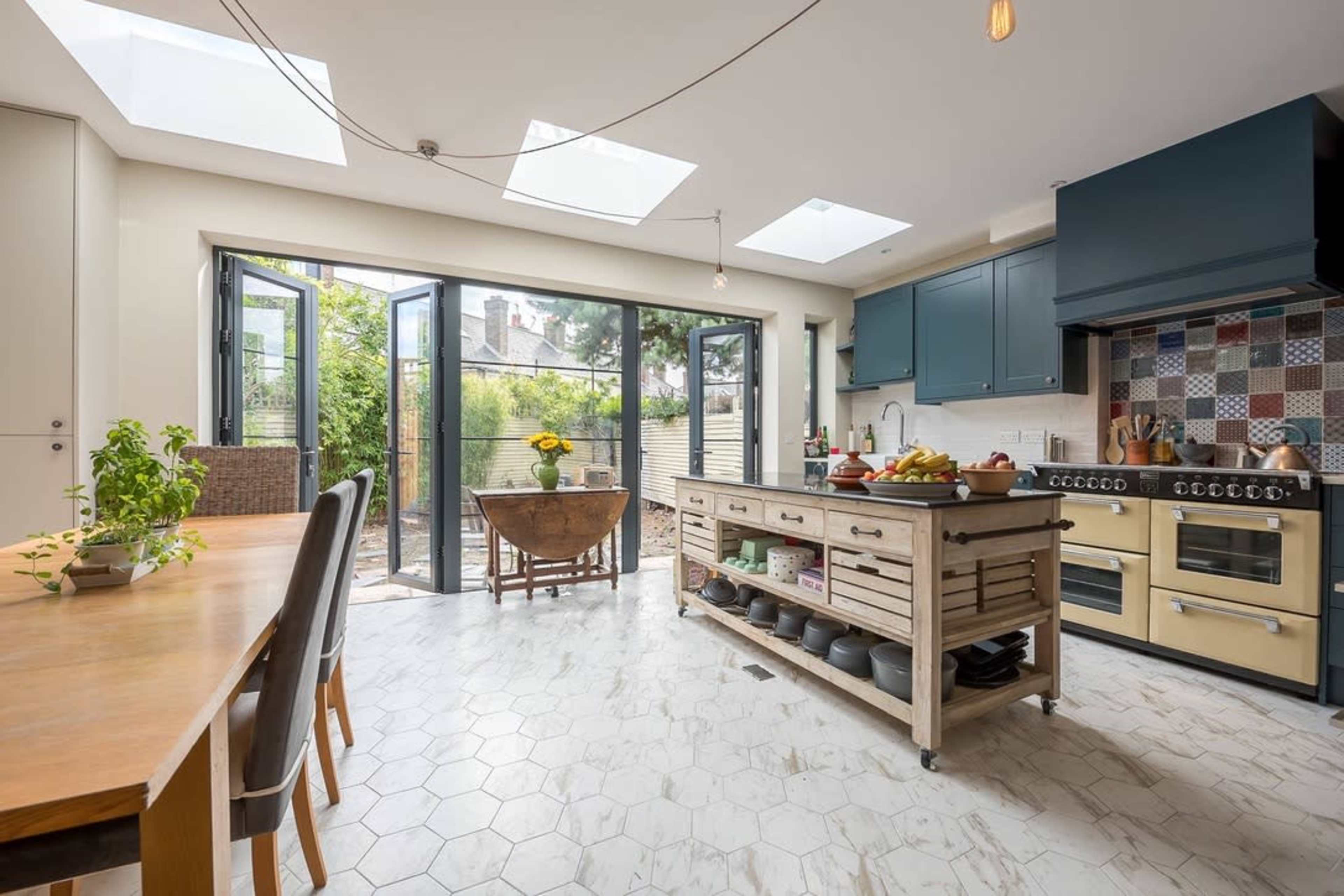 The image shows a modern kitchen with an island, large windows that open to a garden, and a dining table set for meals.