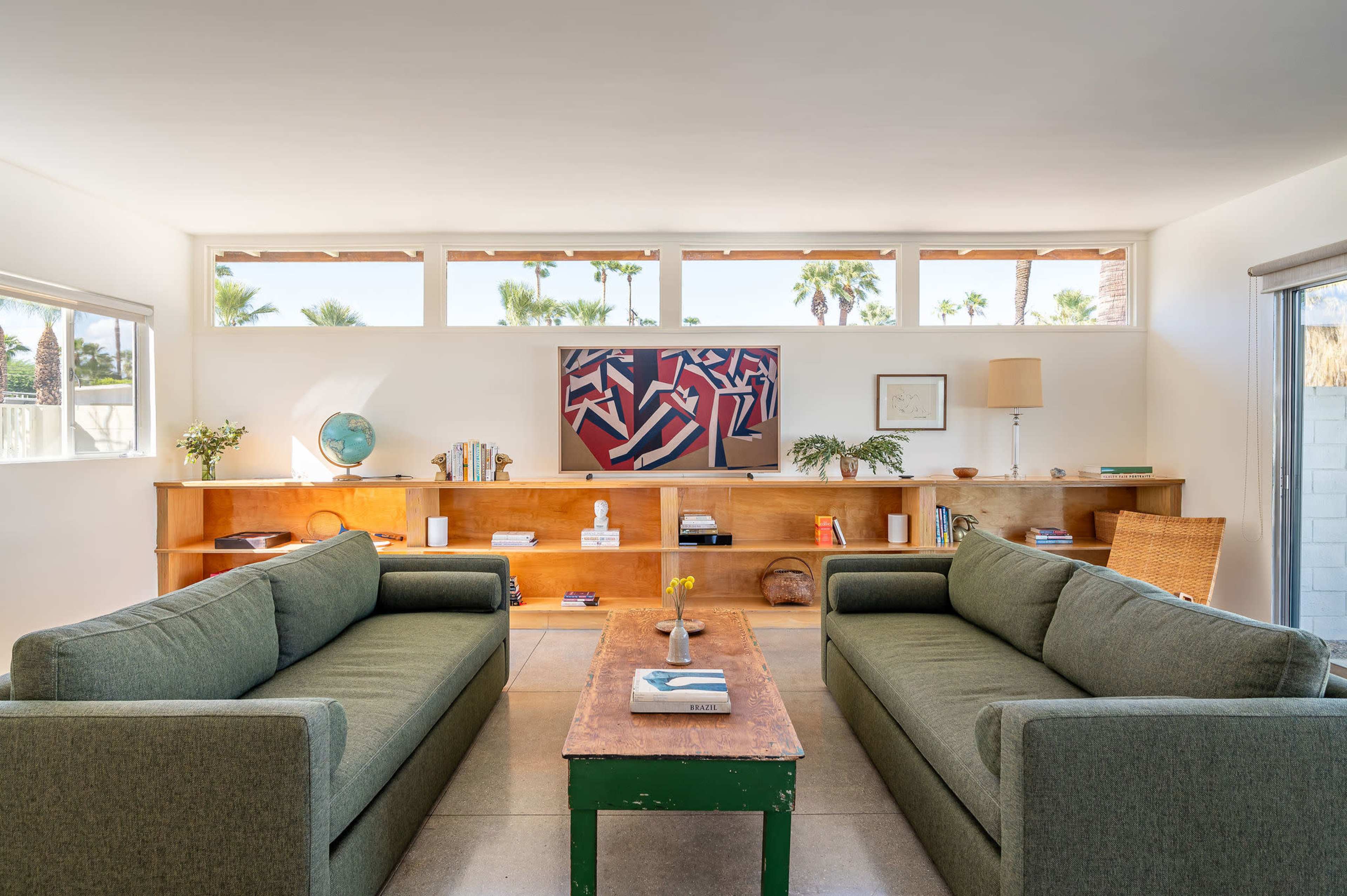 The image shows a modern living room with two green sofas facing a wooden coffee table, a low bookshelf, and large windows providing natural light.