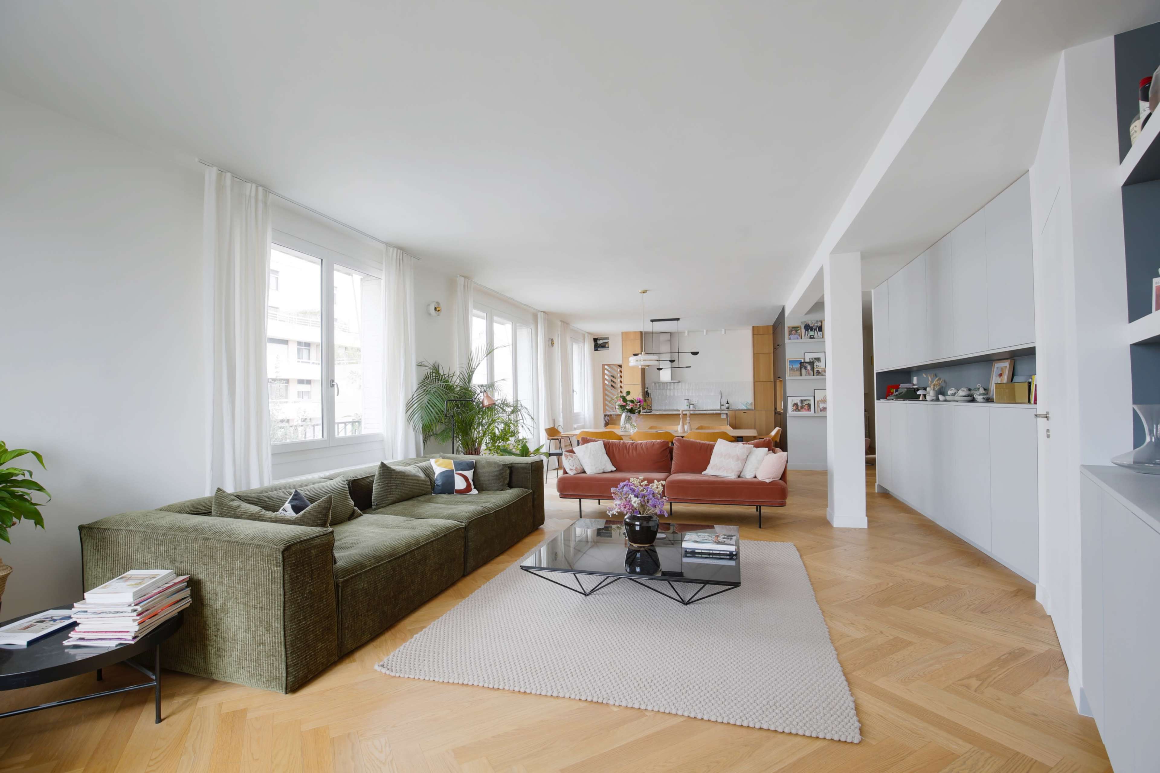 A spacious living room with a green sectional sofa, an orange couch, a coffee table on a light rug, and large windows allowing natural light to enter.