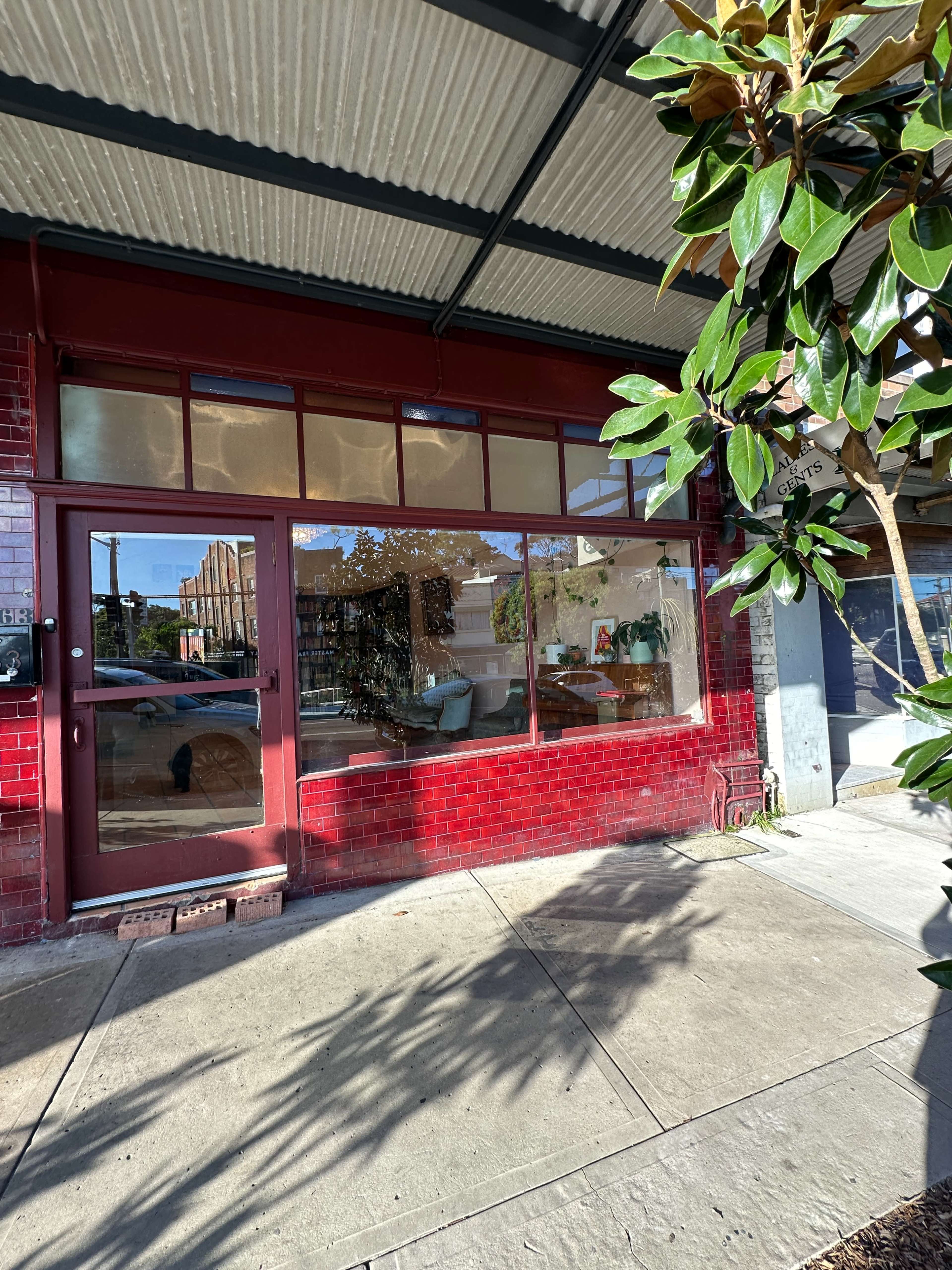 The image shows a storefront with large windows and a red brick facade, partially shaded by a nearby plant.