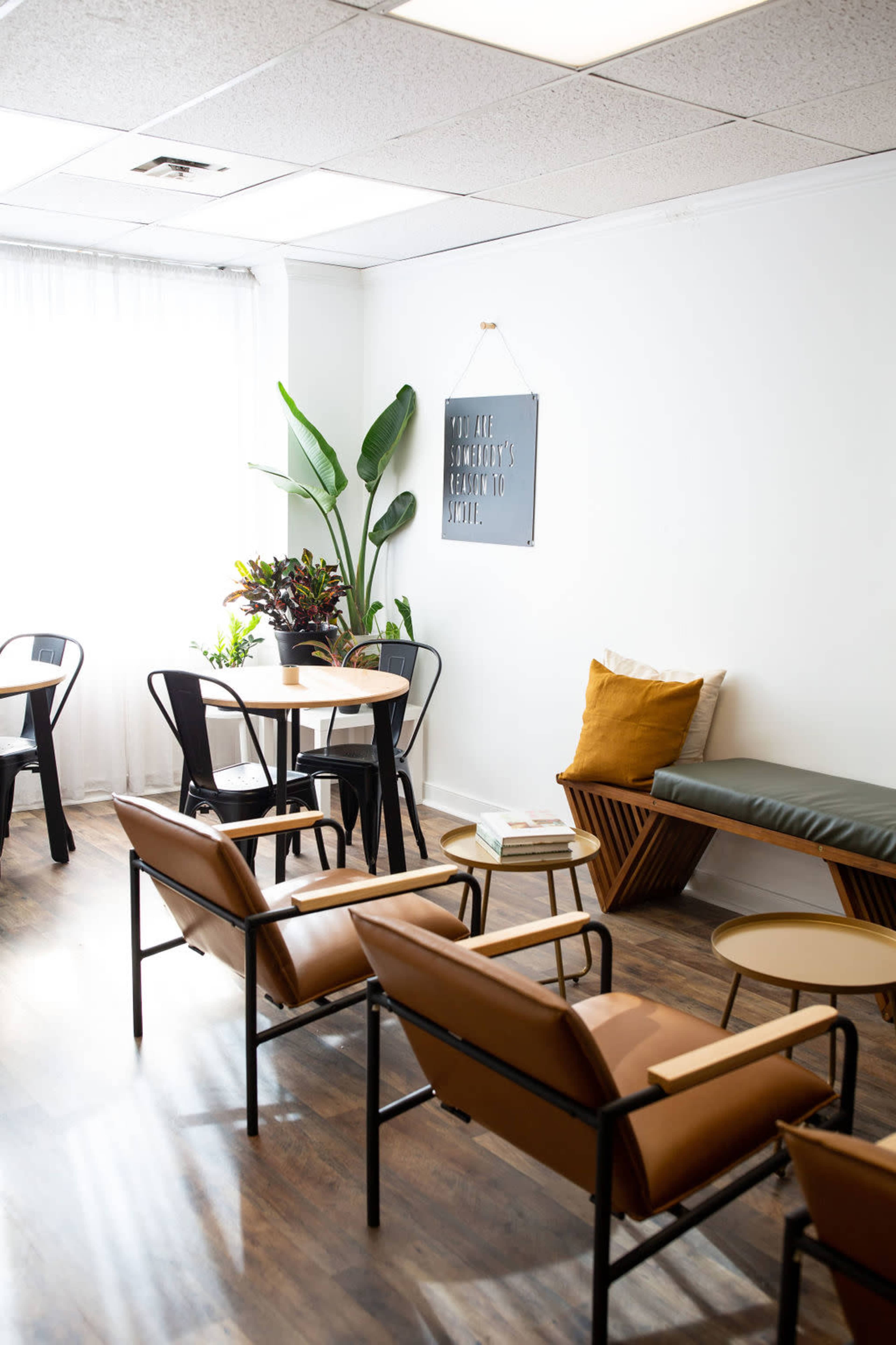 A bright, minimalistic room features wooden flooring, a mix of seating options, a potted plant, and decorative wall art.