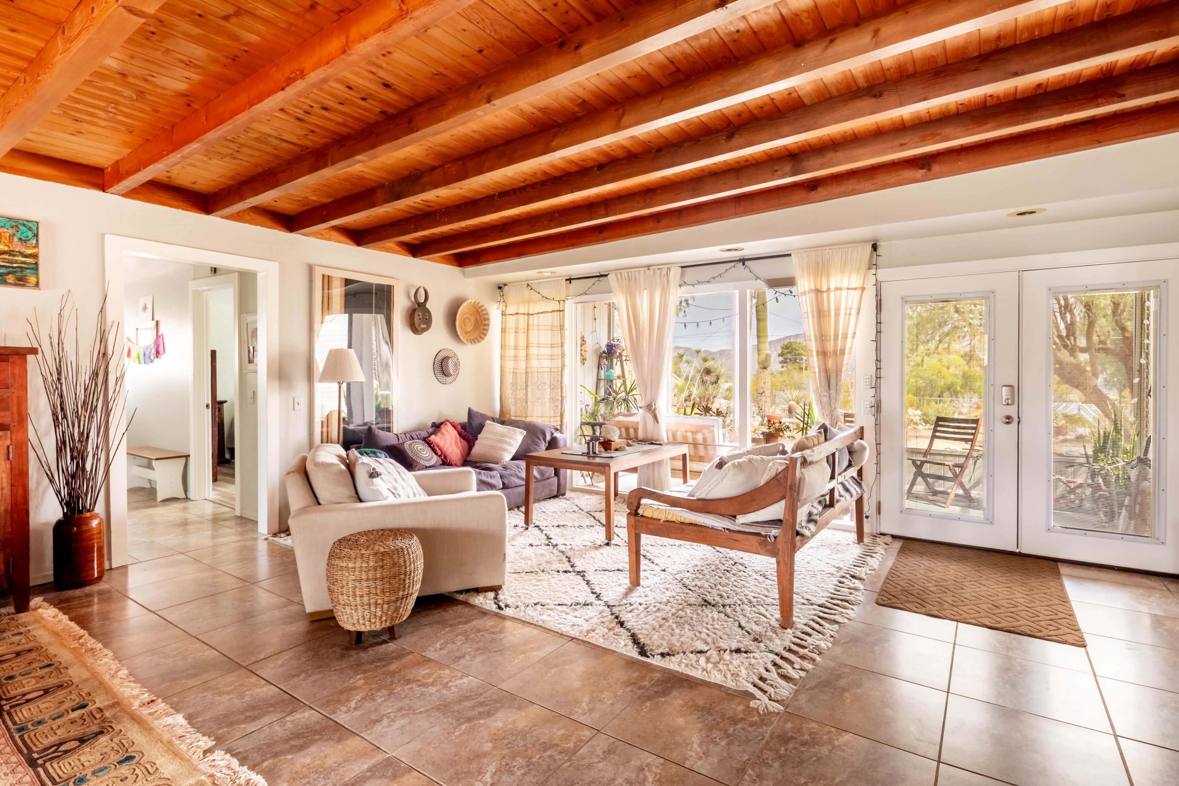 The image shows a bright living room with a wooden ceiling, large windows, and a mix of comfortable seating arrangements inviting natural light.