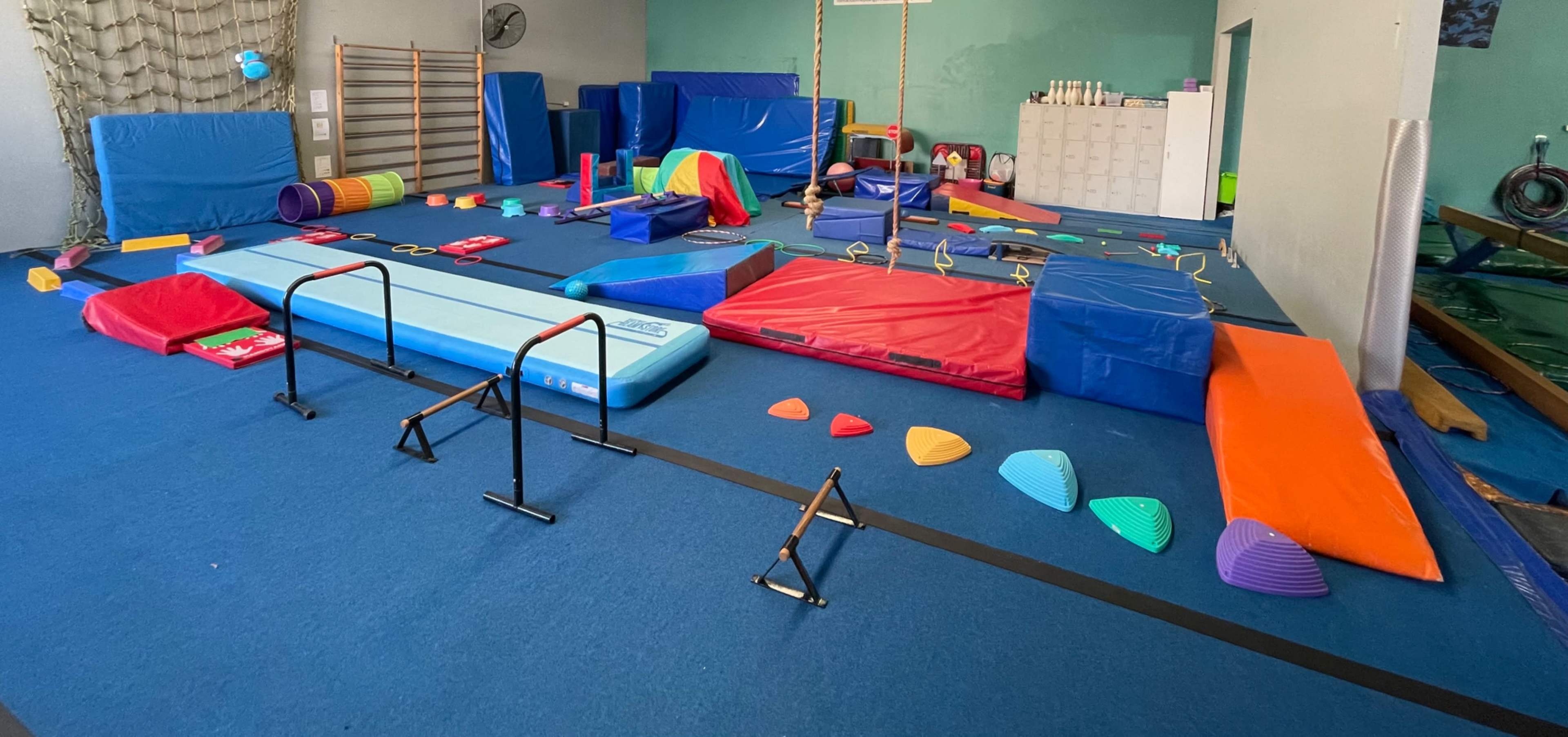A gymnasium with various colorful gymnastic equipment, including mats, balance beams, and obstacle course elements, arranged across the blue floor.