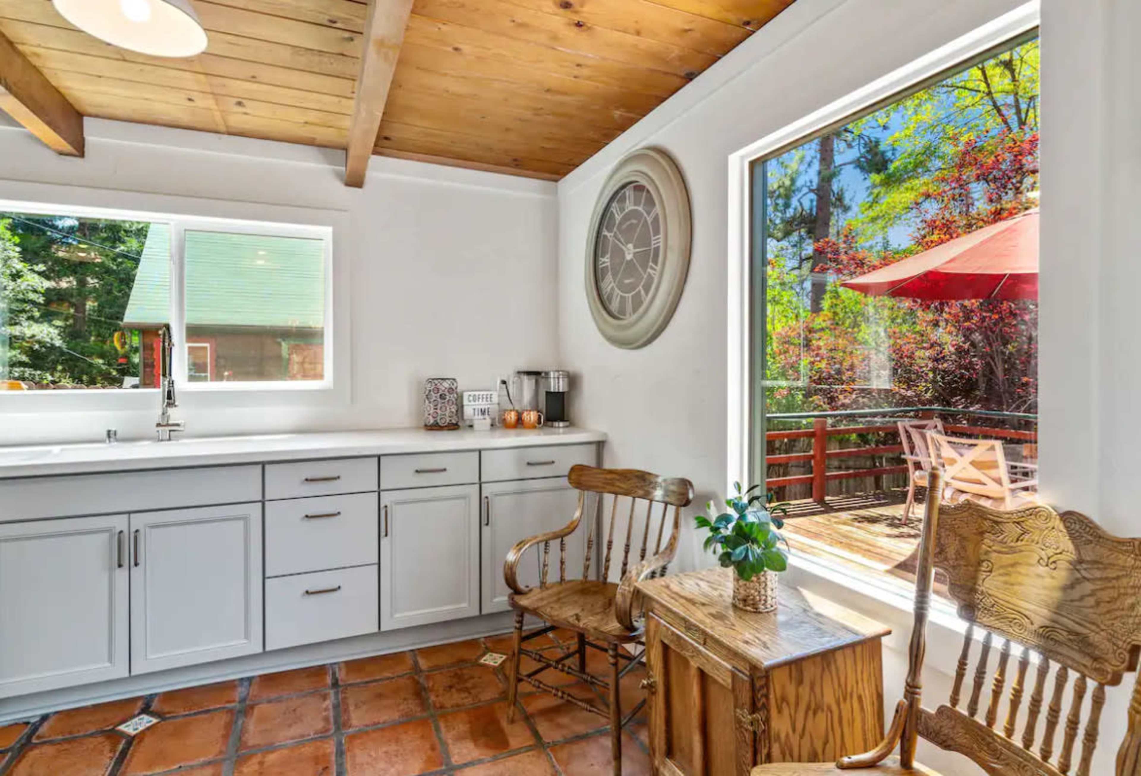 A bright kitchen features white cabinets, a wooden ceiling, a clock on the wall, and a view of an outdoor deck and trees through large windows.