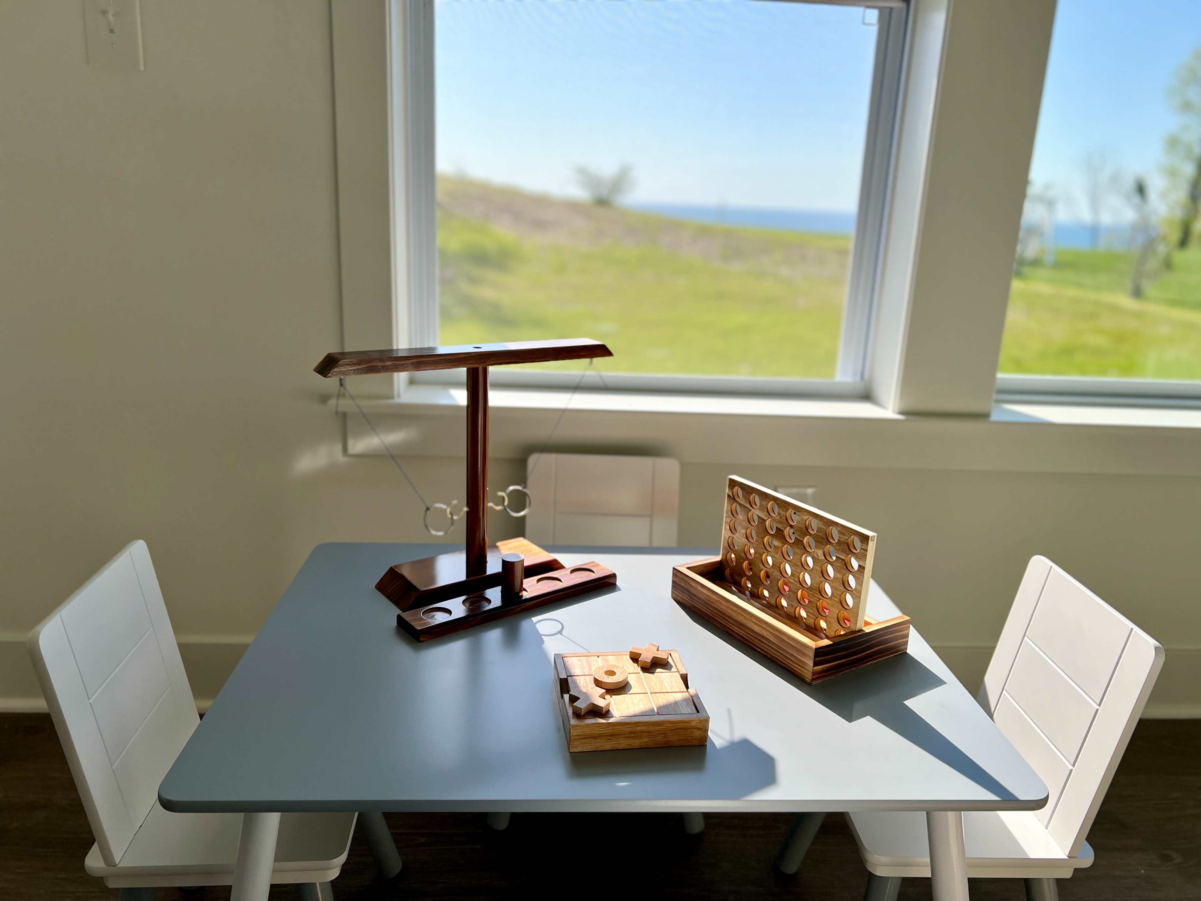 A wooden activity table set up with three games is positioned near a window overlooking a grassy area and water in the background.