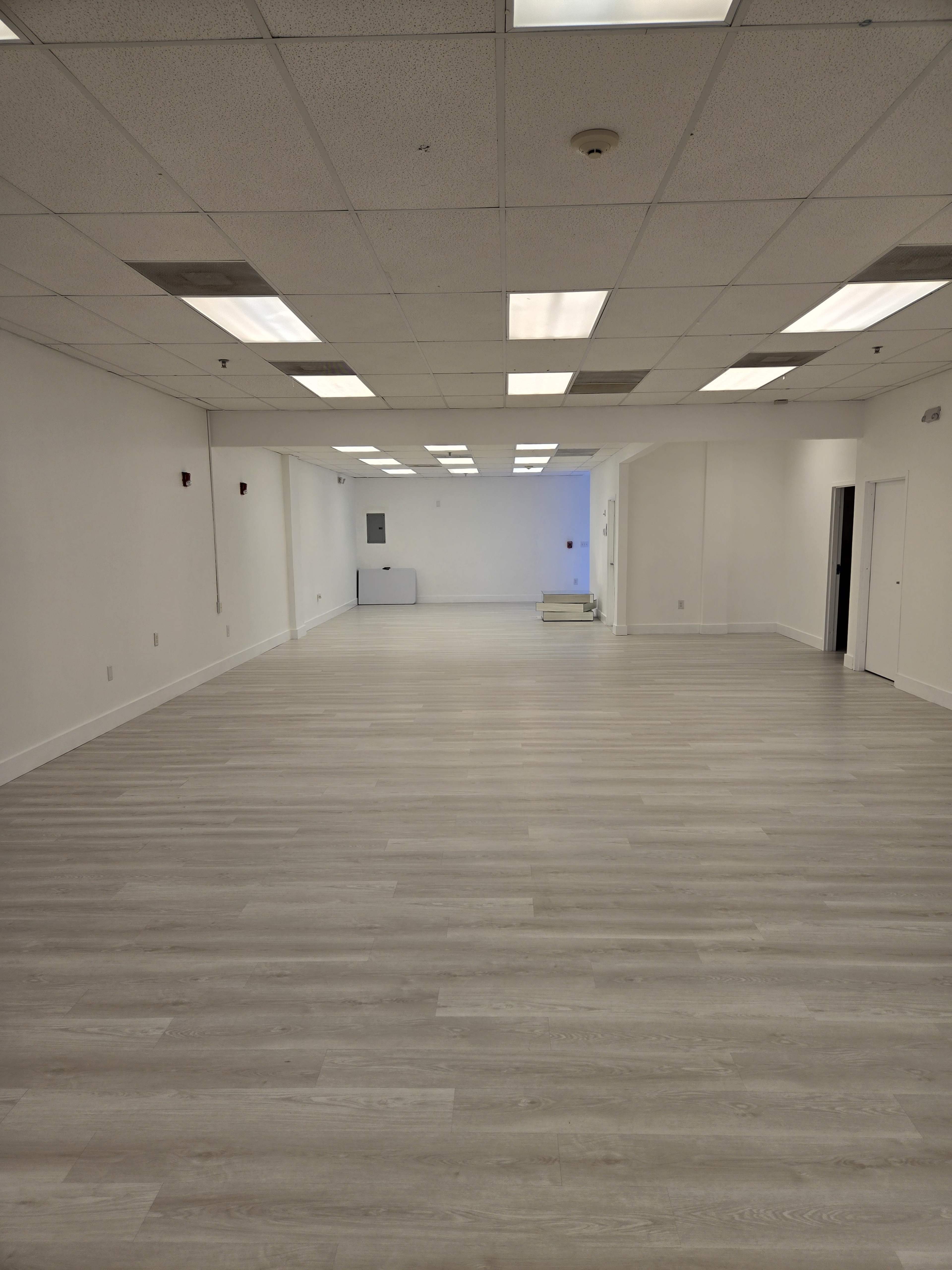 A spacious, empty room with light wood flooring and overhead fluorescent lighting.