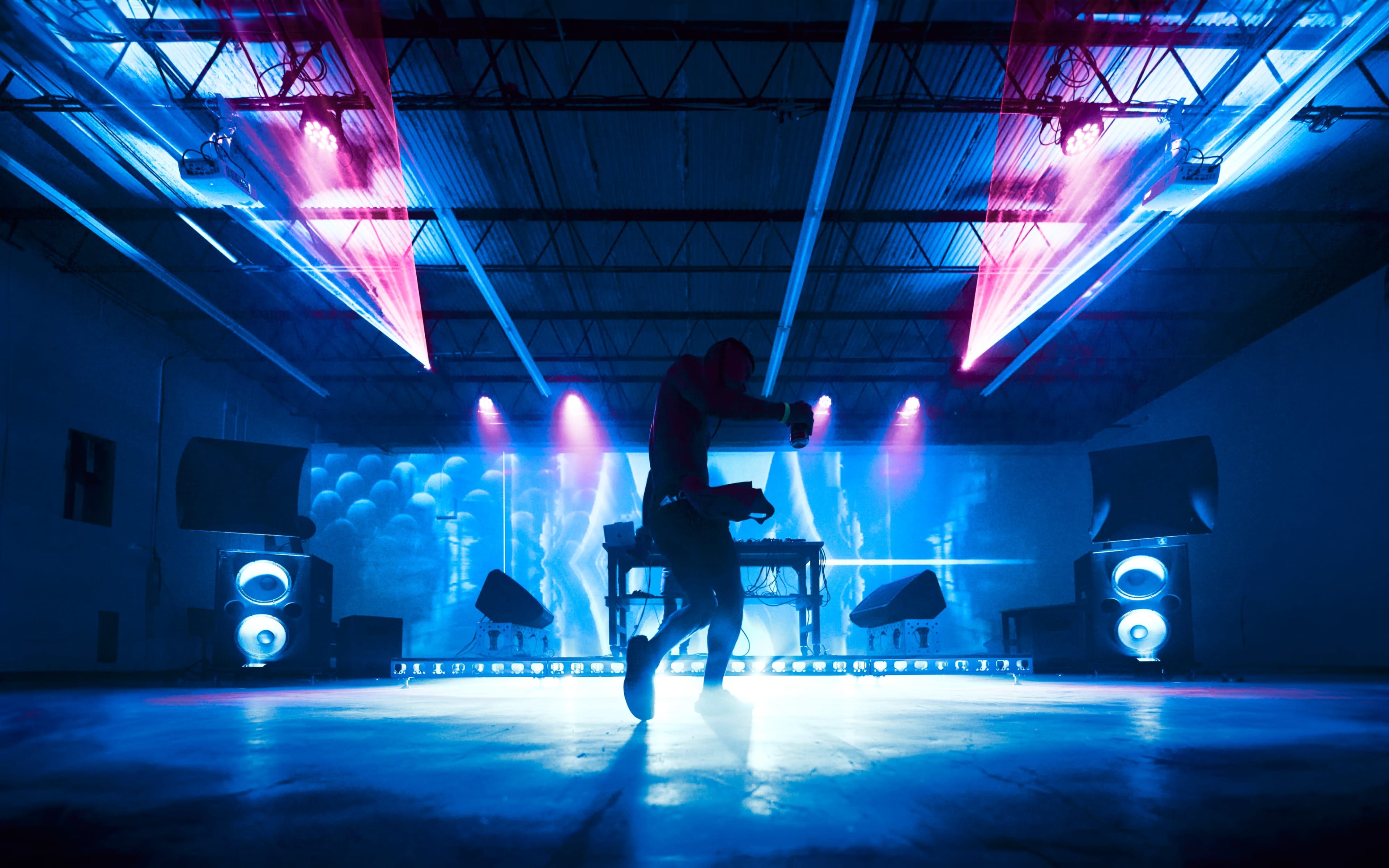 A dancer performs on stage in a dimly lit industrial space with colorful lights and a DJ setup in the background.