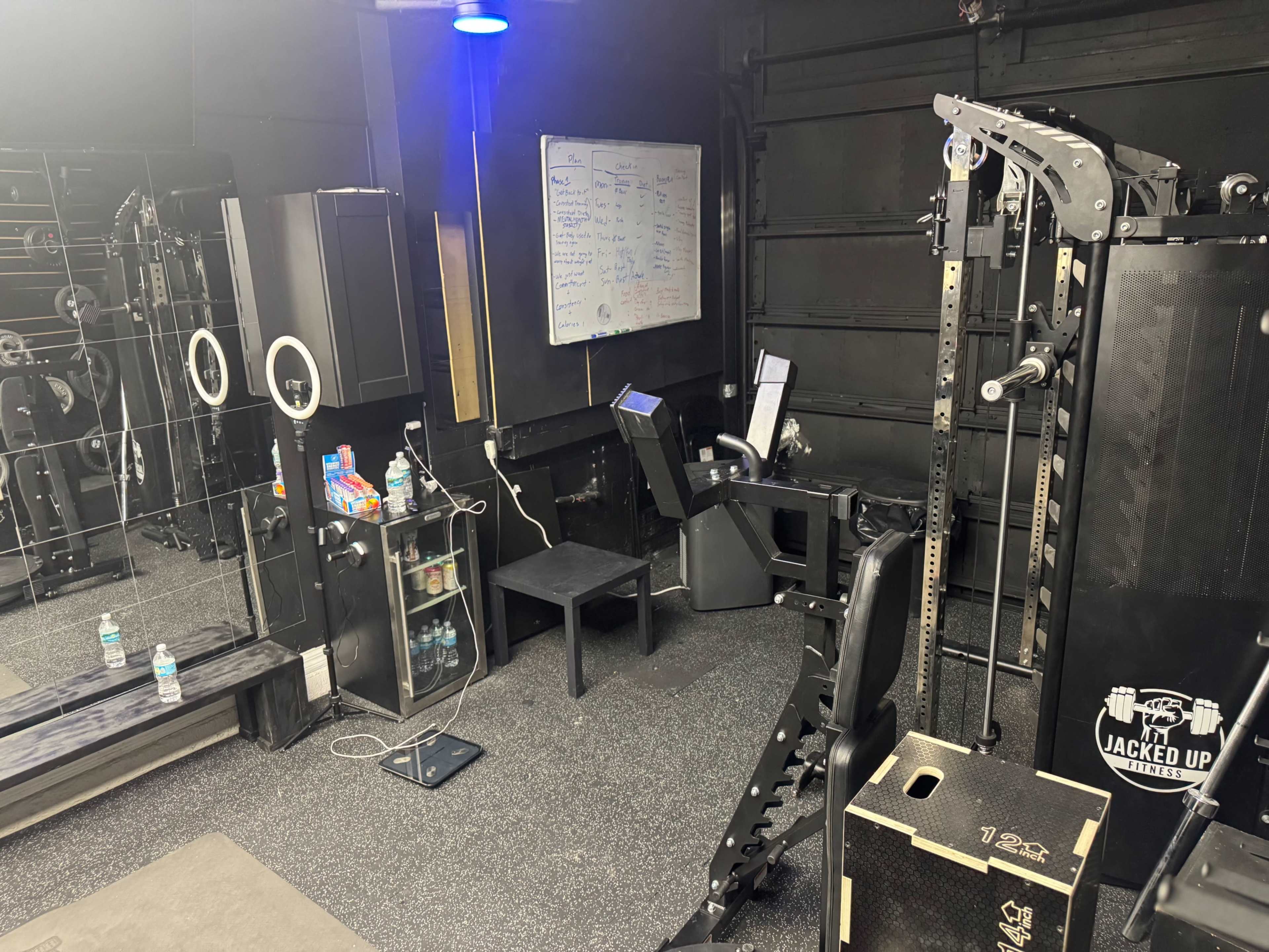 The image shows a home gym setup featuring a power rack, a mirrored wall, a chalkboard, and several fitness equipment and water bottles on a black rubber floor.