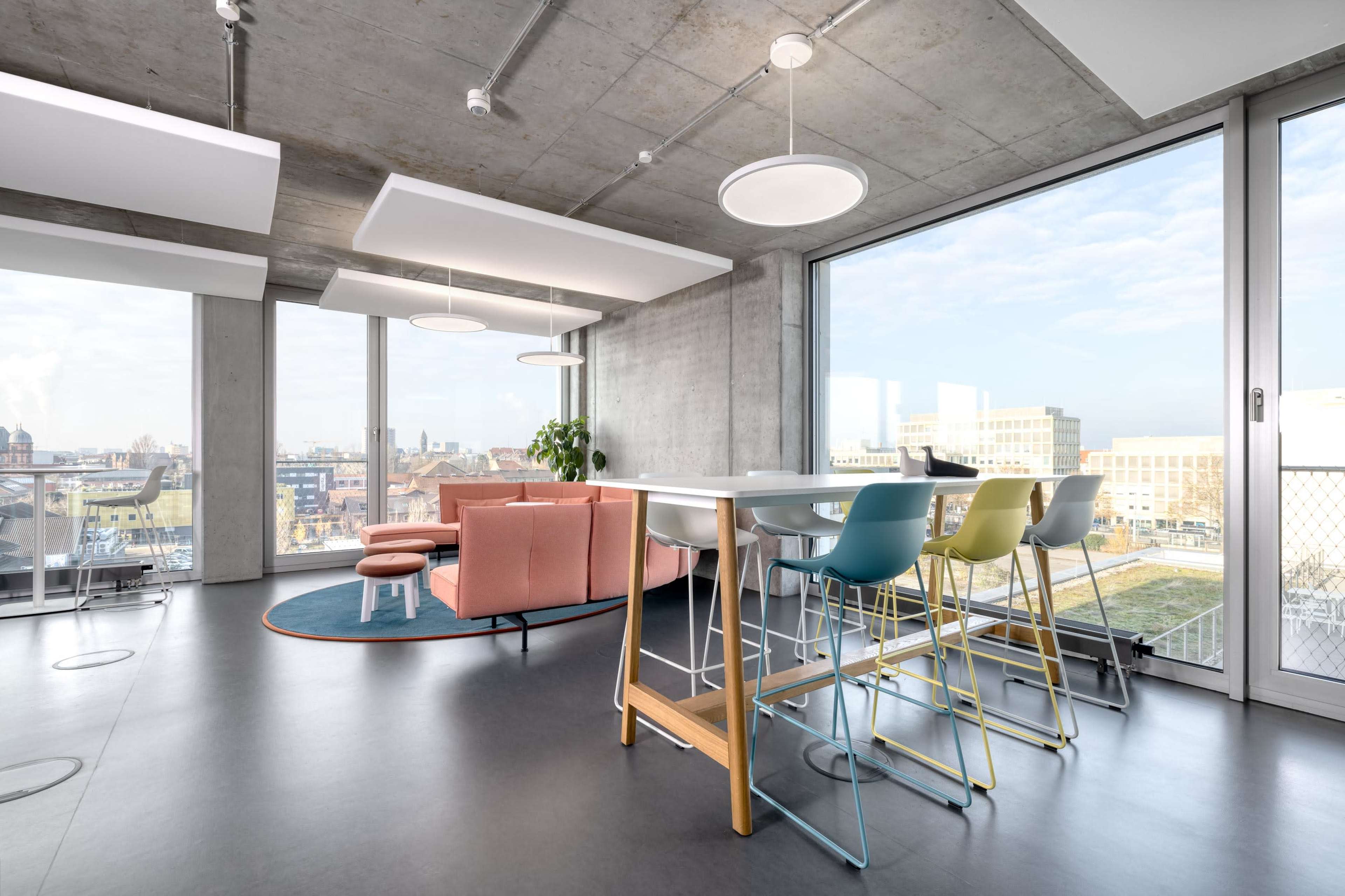 A modern office space with a pink sofa and a dining area featuring colorful bar stools, large windows, and a view of the cityscape outside.
