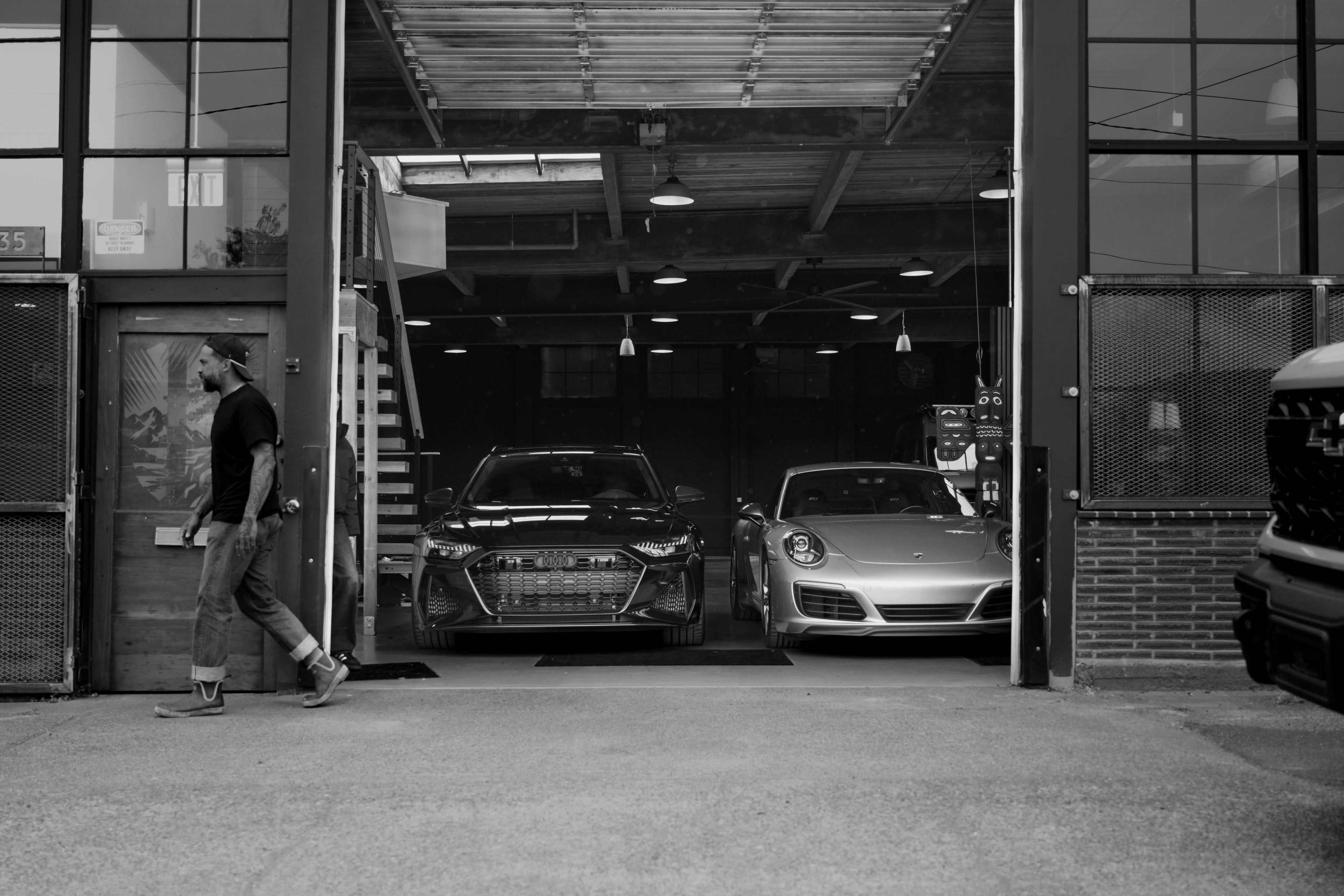 A man walks past two luxury cars parked inside a spacious garage bay with large open doors.