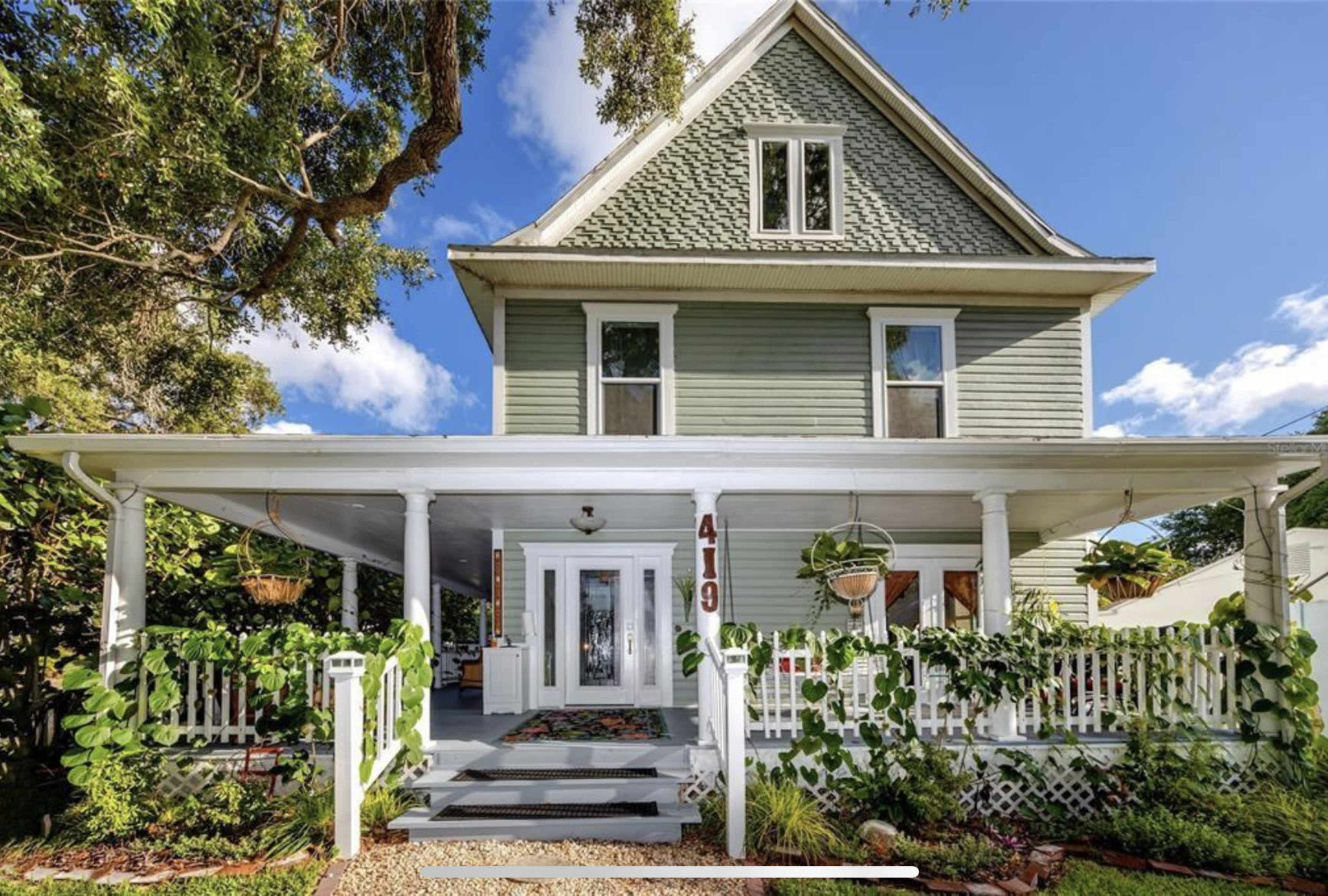 The image shows a large, two-story green house with a front porch adorned with plants and a white picket fence.