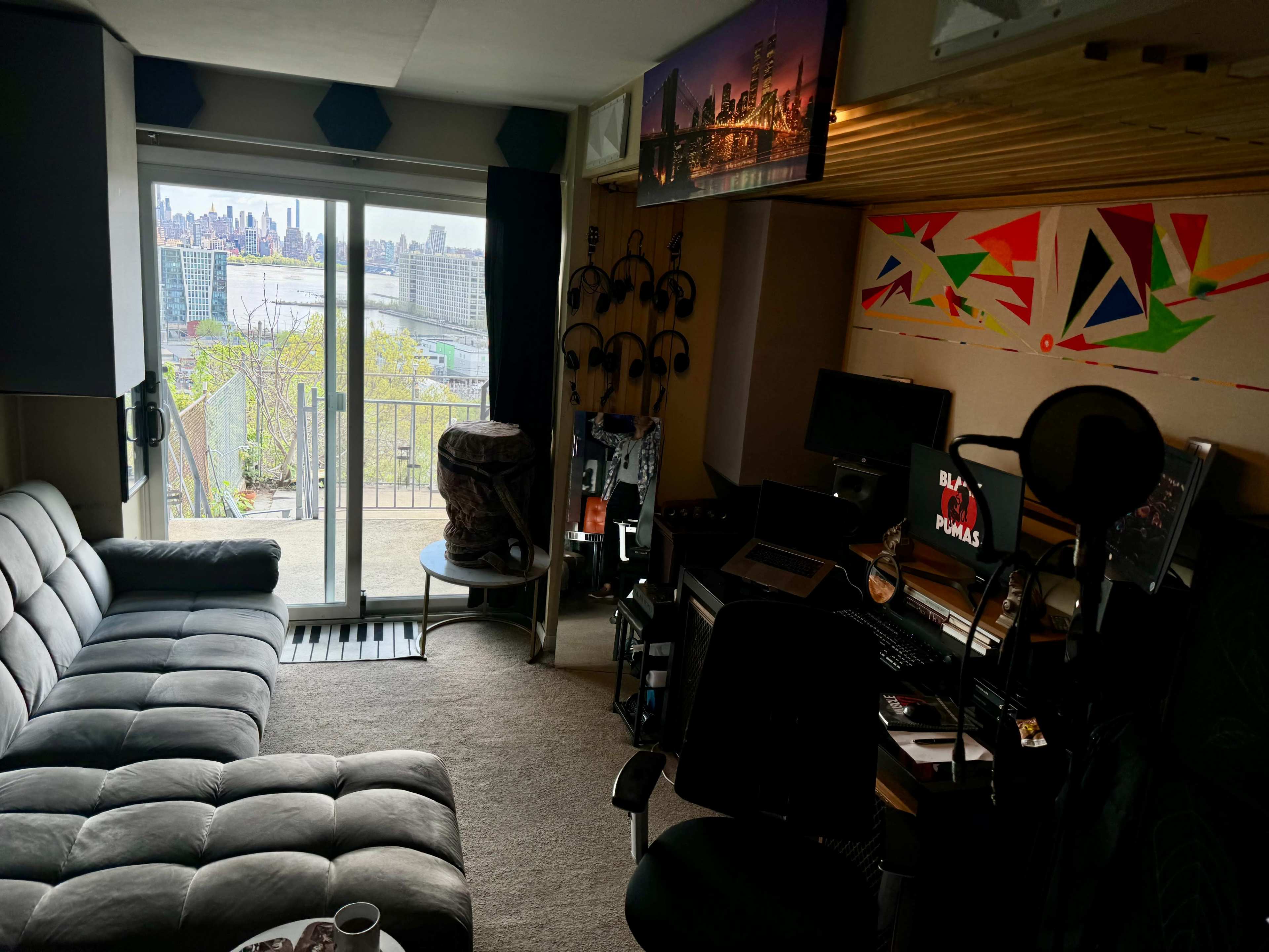 The image shows a cozy room featuring a sofa, a desk with equipment, and a view of a city skyline through sliding glass doors.
