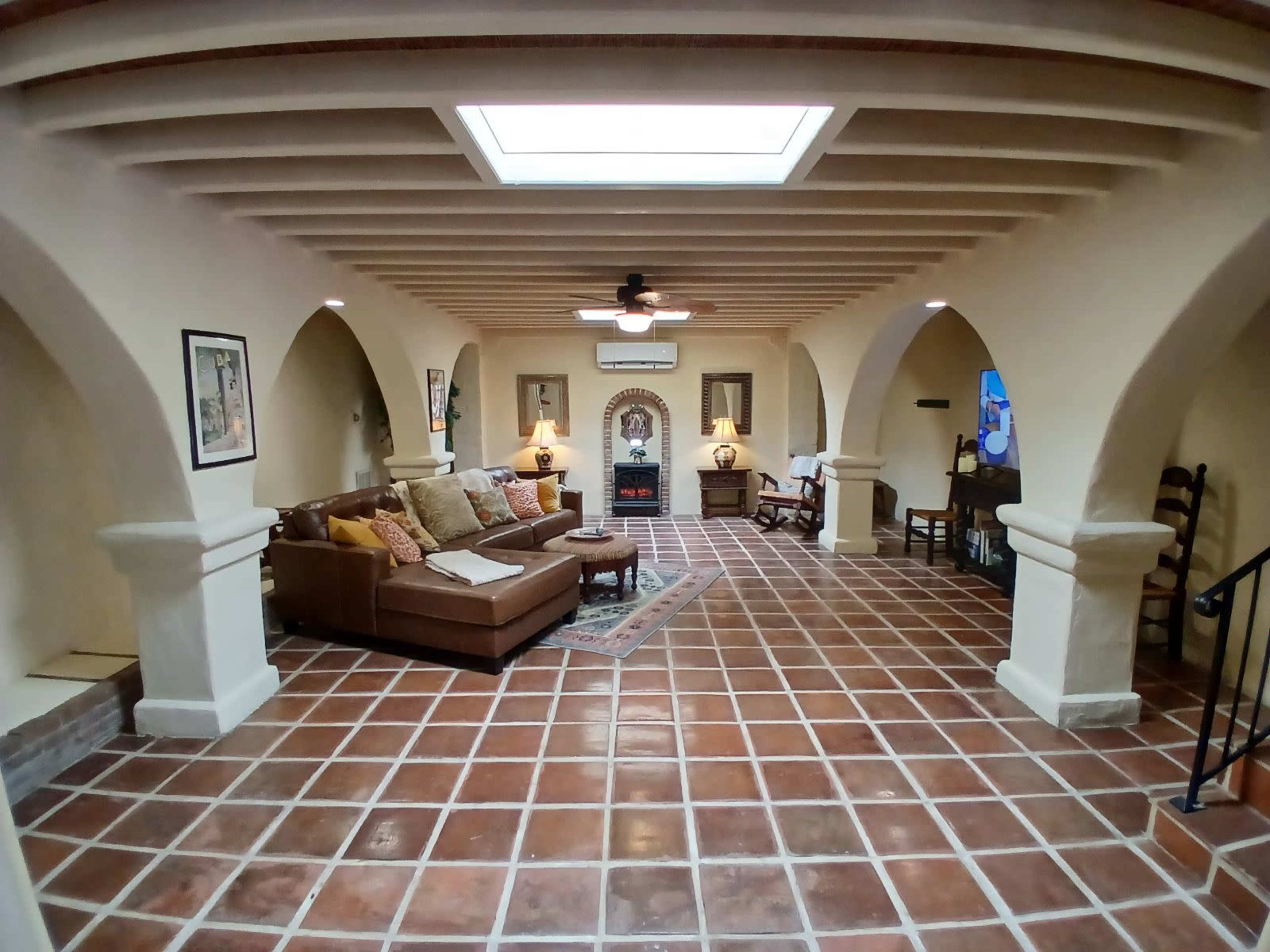 A spacious, well-lit living room with arched openings, a brown sofa, a decorative rug, and a fireplace, featuring tile flooring and ceiling beams.