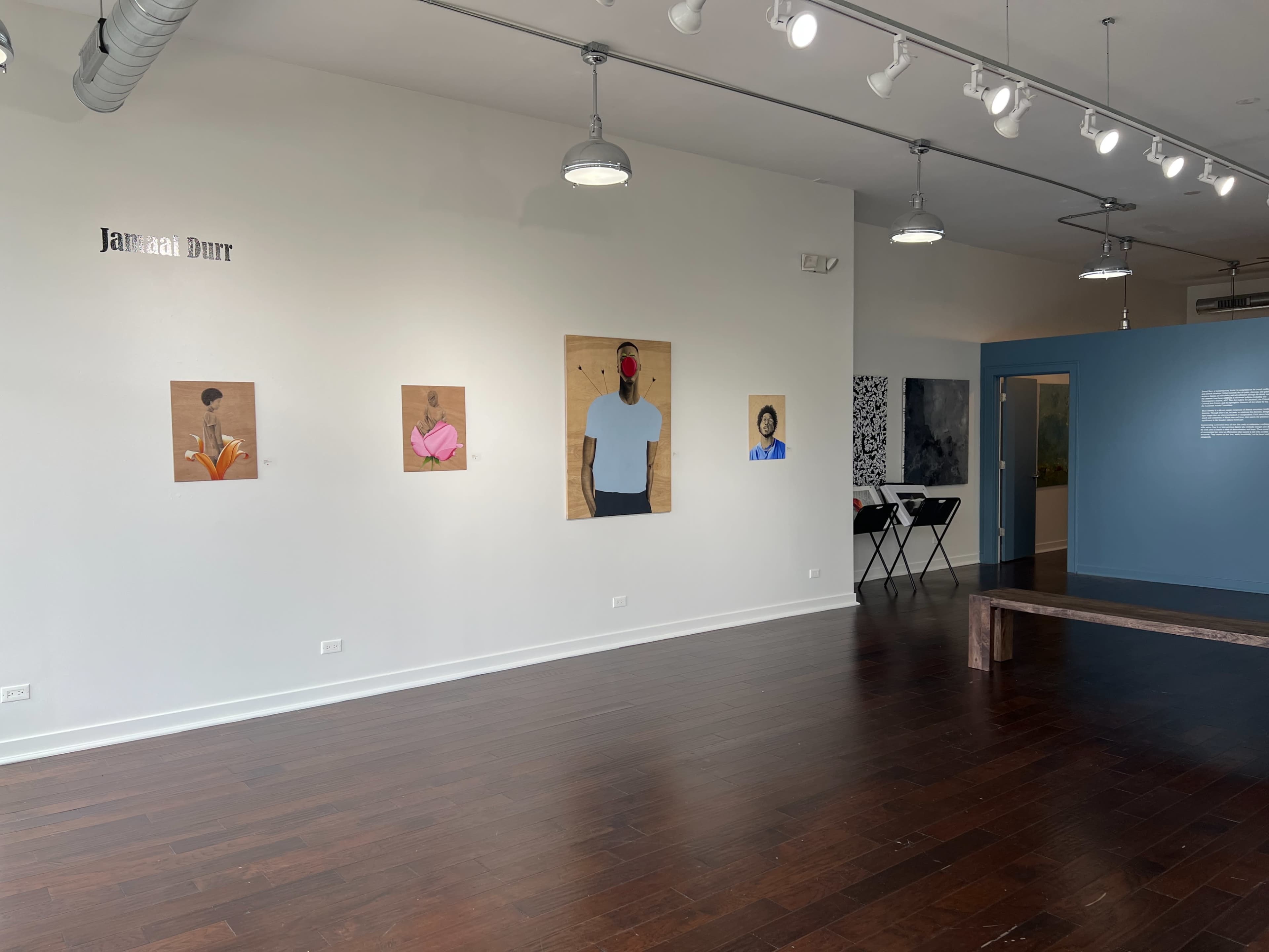 An art gallery with a series of framed paintings hung on a white wall, featuring portraits of various people, while a blue wall is visible in the background.
