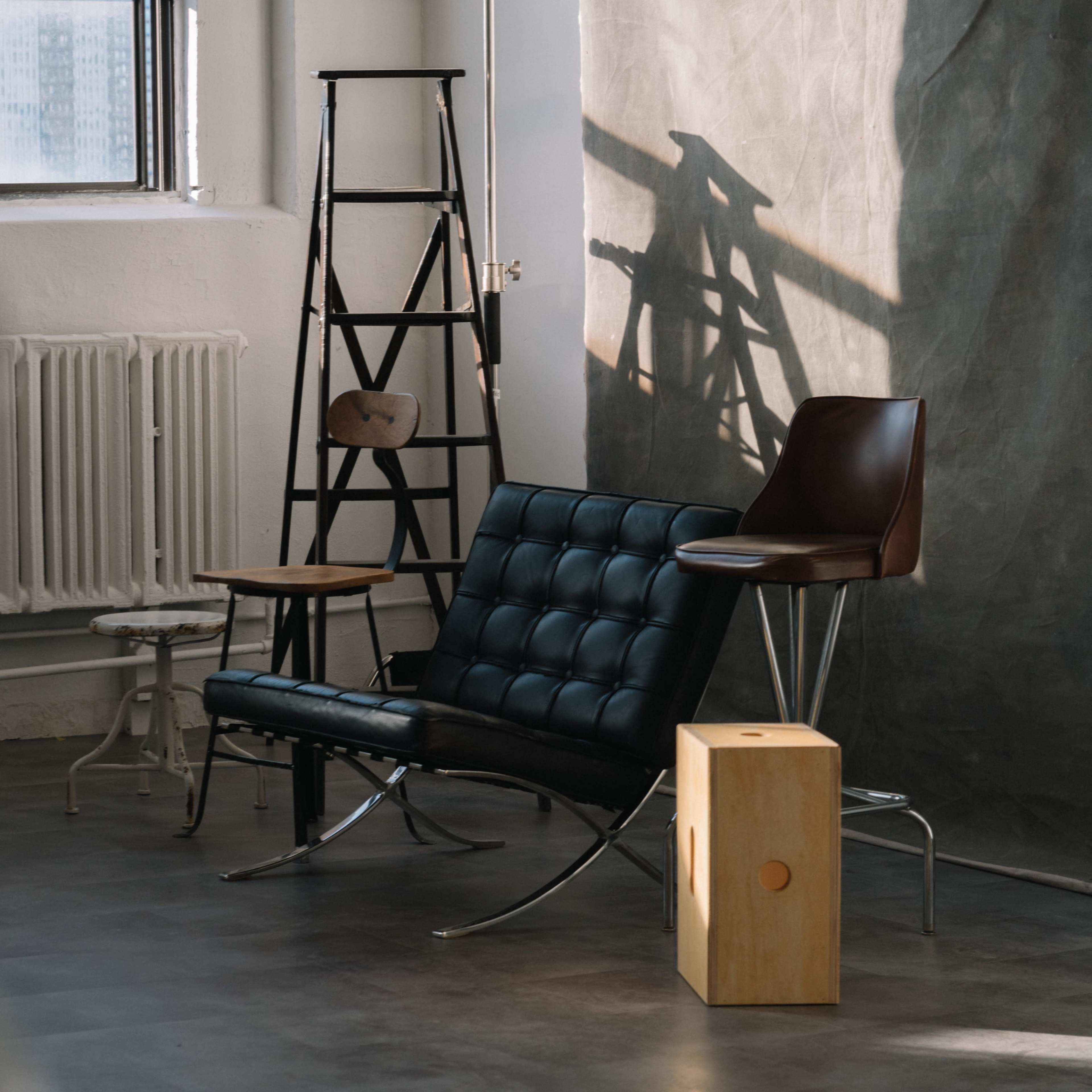 A modern interior setting with a black leather sofa, a wooden stool, a high brown chair, a wooden block, and a metal ladder against a textured wall.