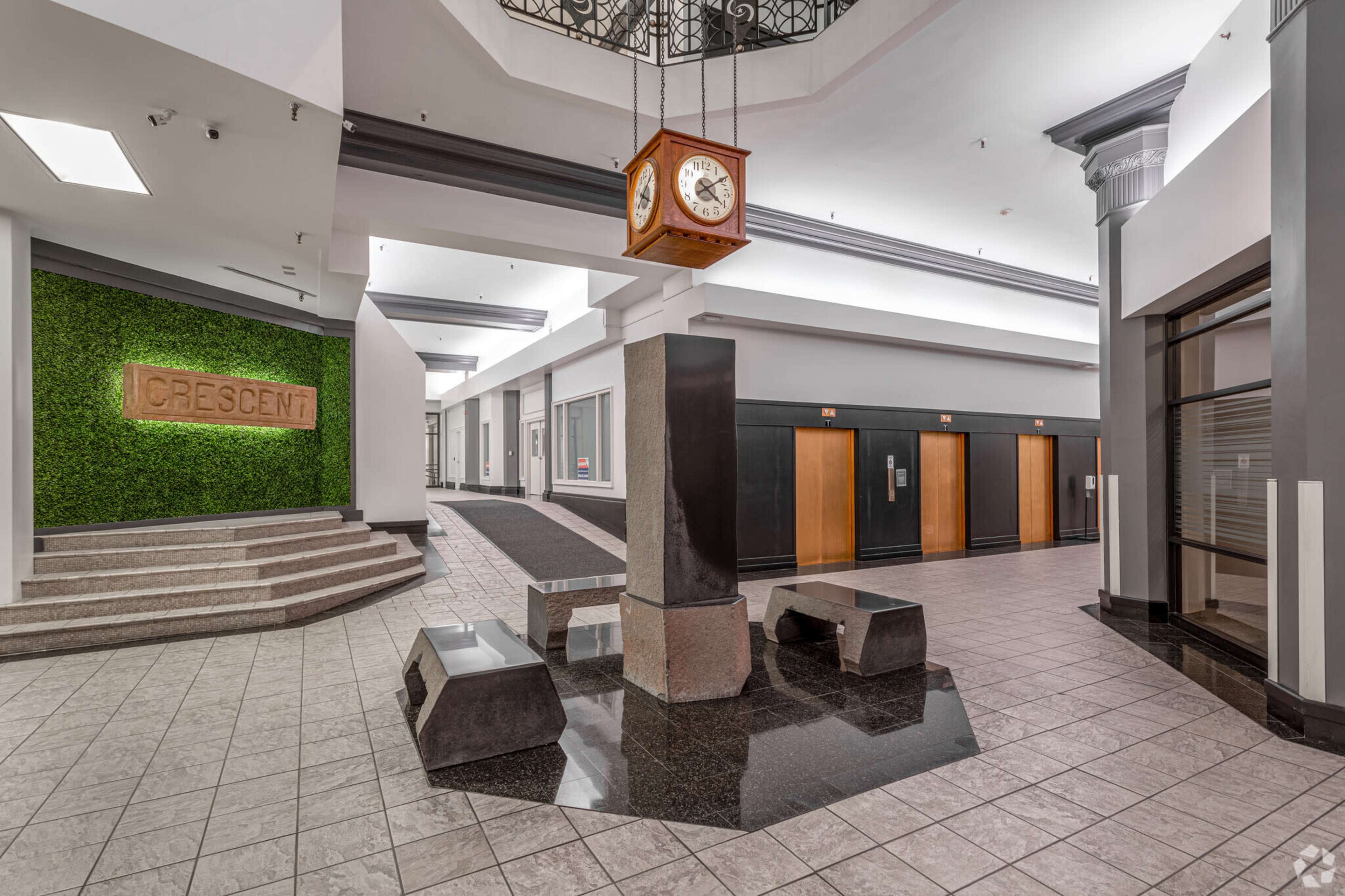 A modern interior lobby with a clock suspended from the ceiling, a decorative green wall, and elevator doors on two sides.