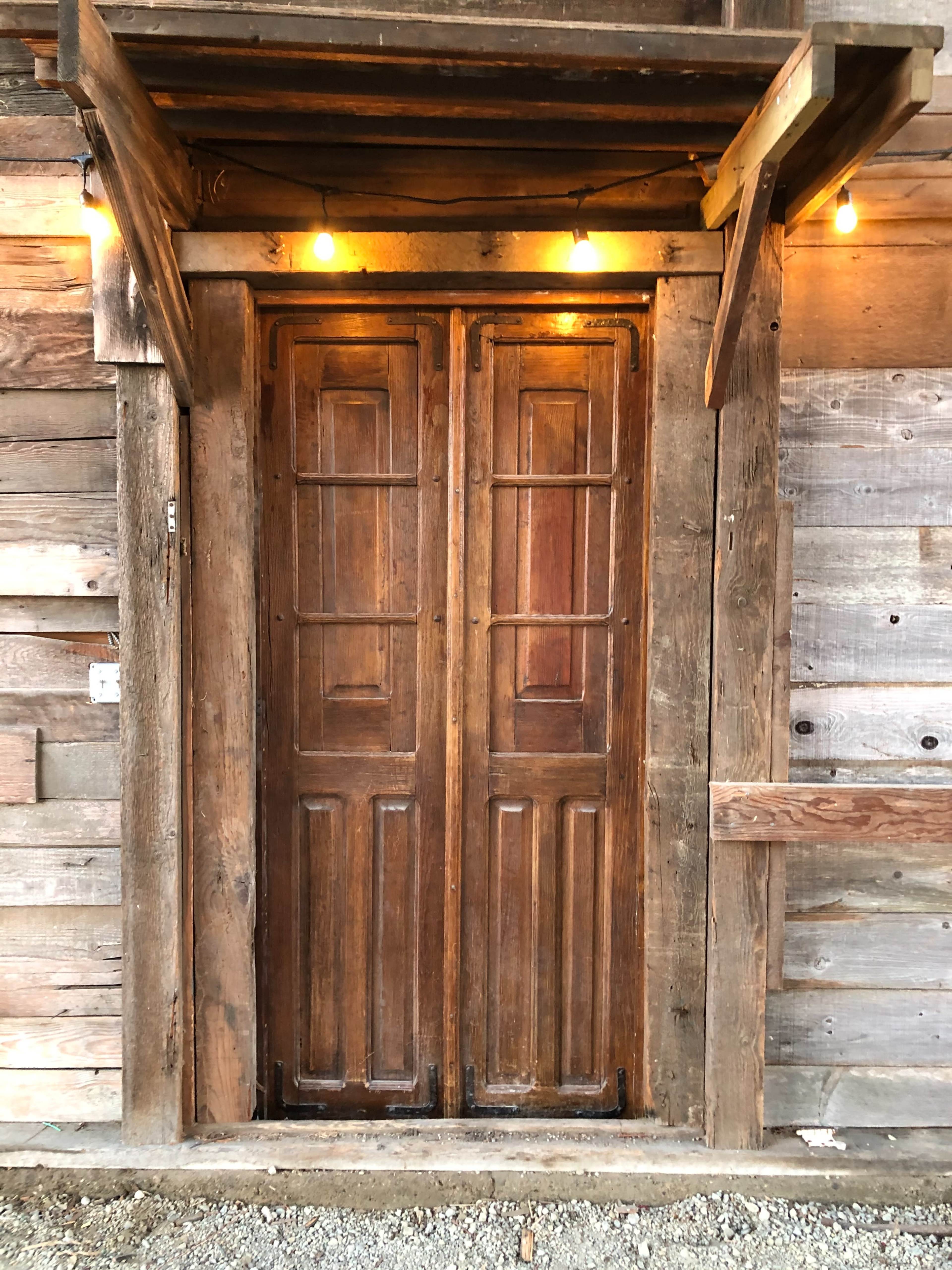 A set of wooden double doors is illuminated by overhead string lights against a backdrop of rustic wooden planks.