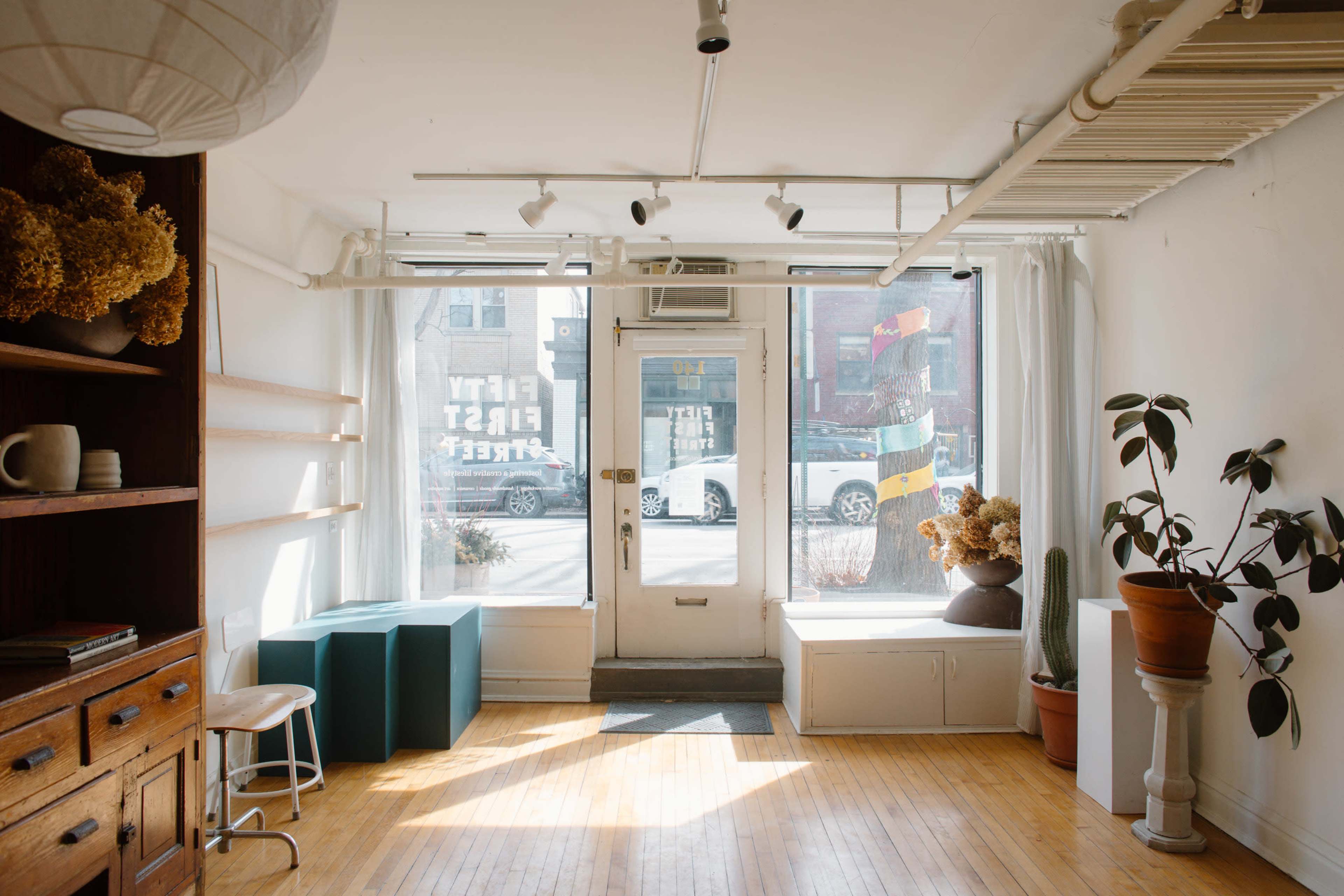 The image depicts a well-lit interior space with wooden flooring, featuring a seating area, potted plants, and large windows that provide a view of the street outside.