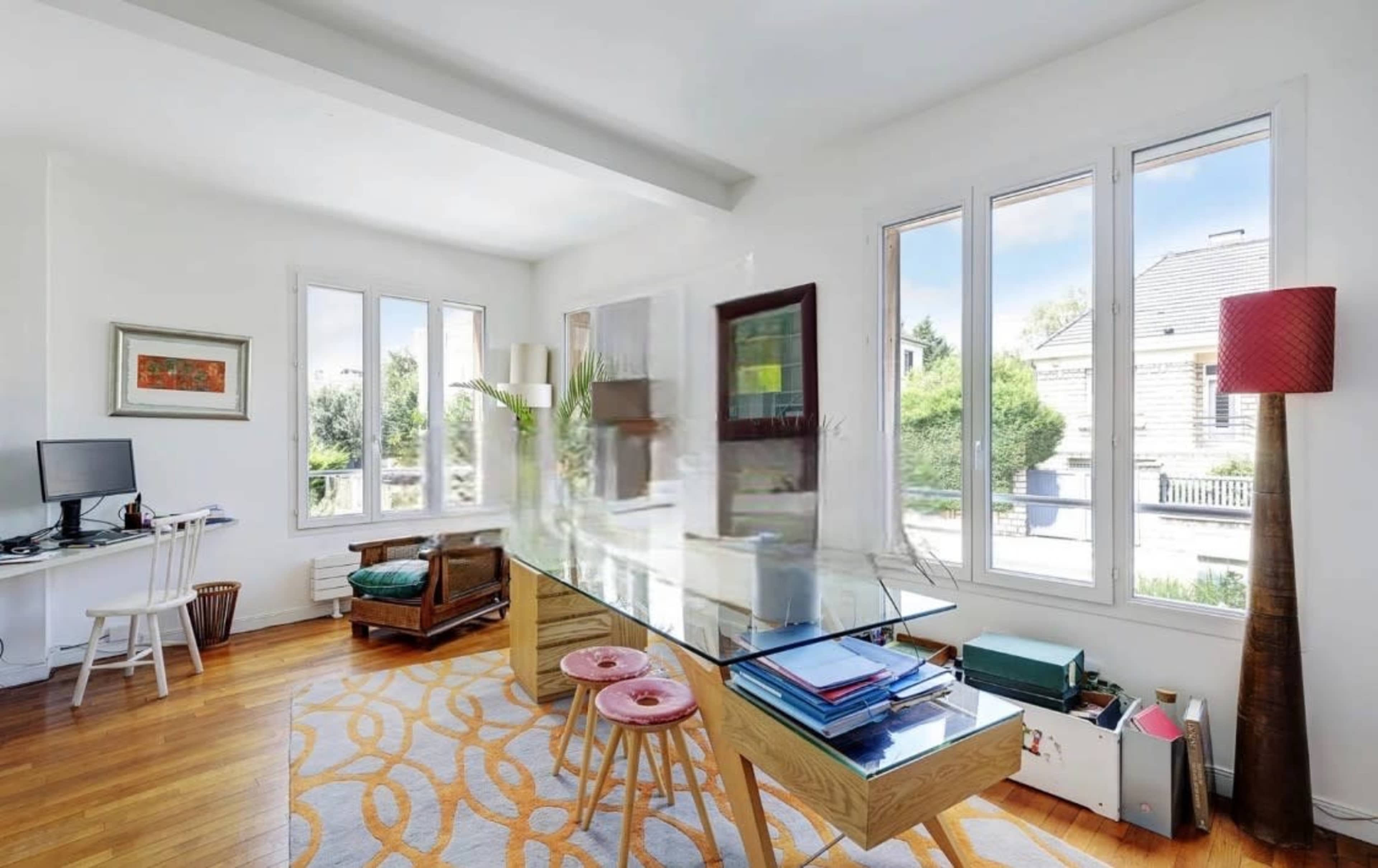 The image shows a bright, modern office space with a glass desk, a couple of pink stools, and large windows that allow natural light to fill the room.