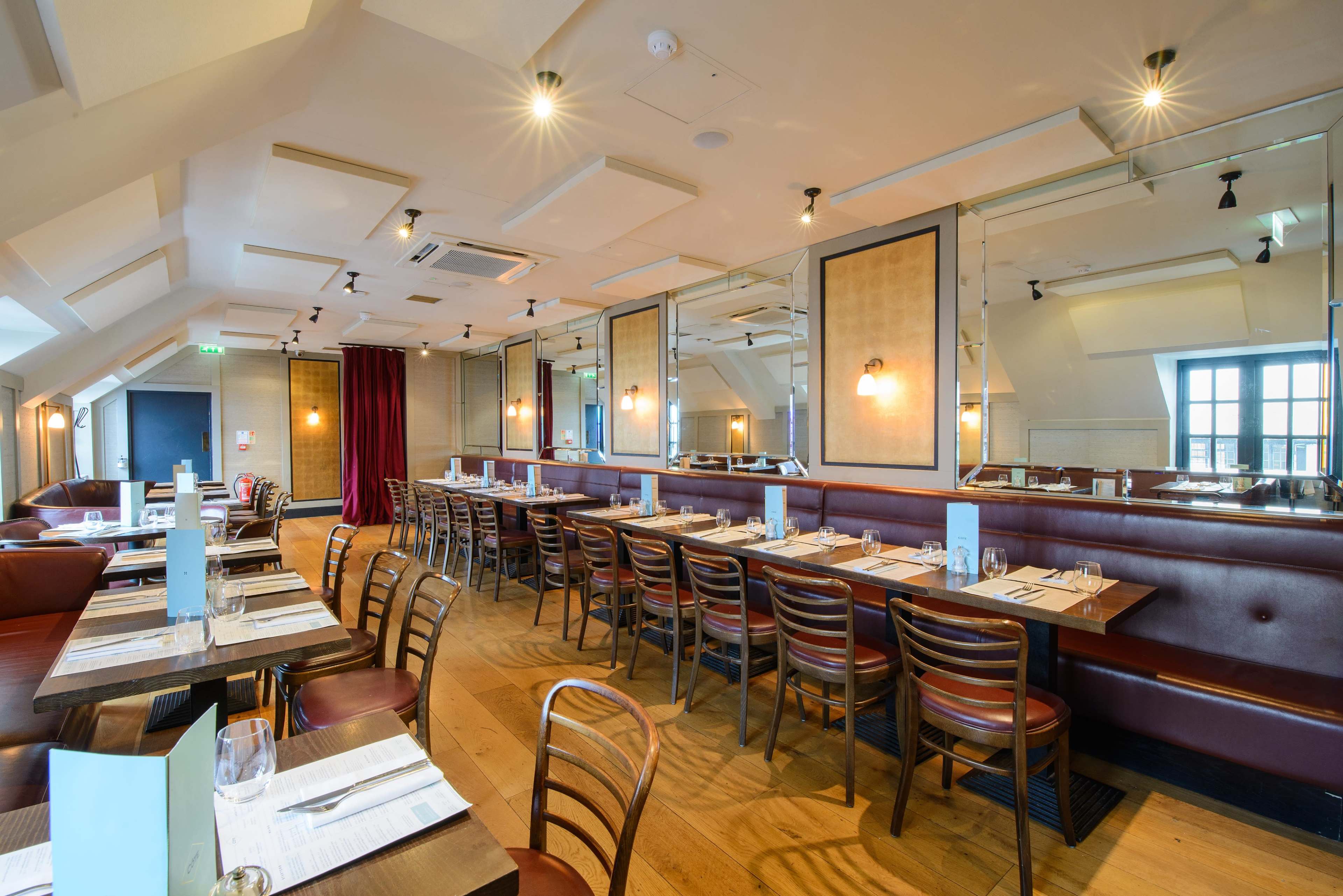 The image shows a modern restaurant interior with wooden flooring, a row of tables set for dining, and mirrors on the walls.