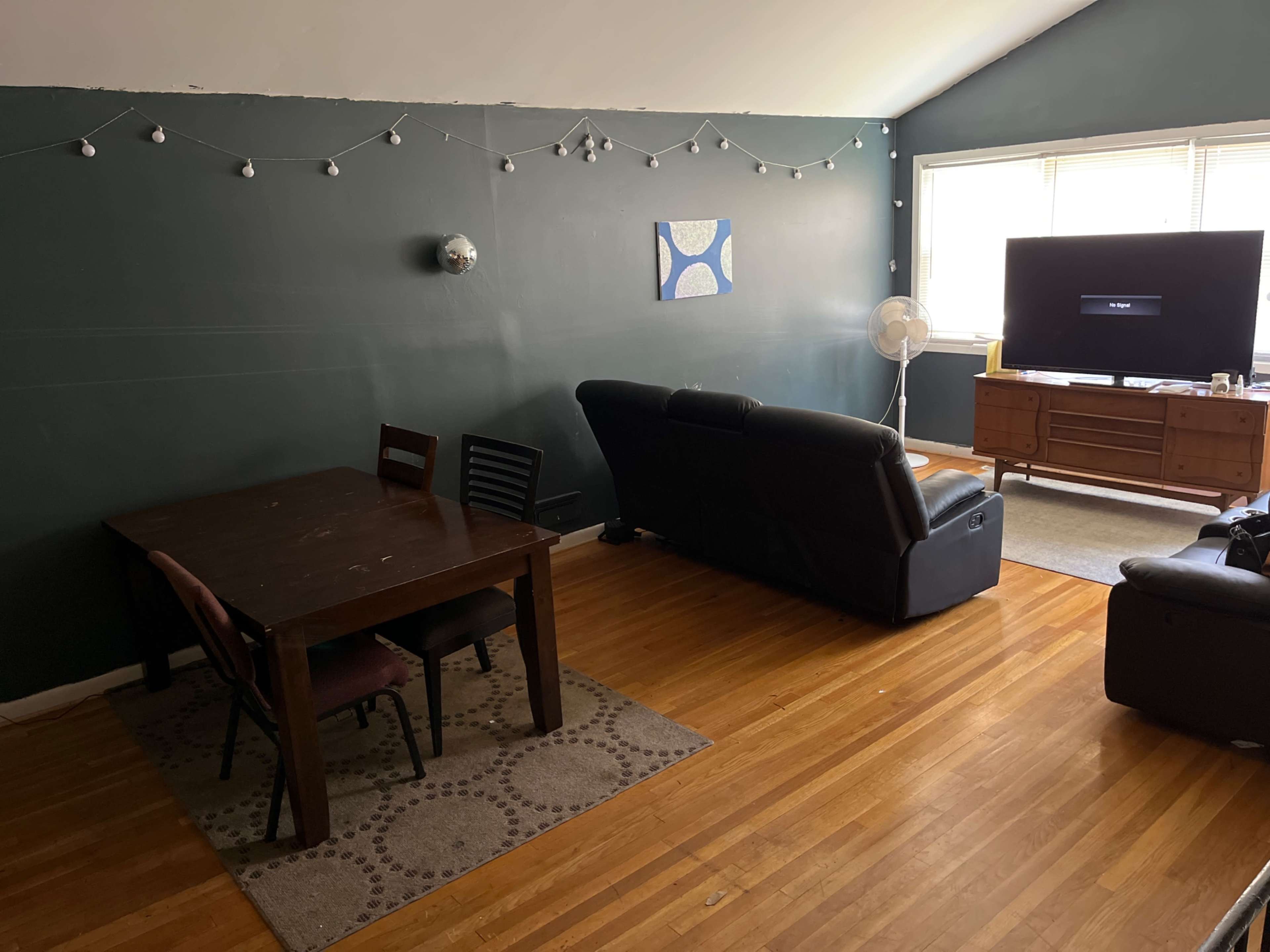 A living room features a seating area with a couch and a television, alongside a dining table and chairs in a space with dark green walls and wooden flooring.