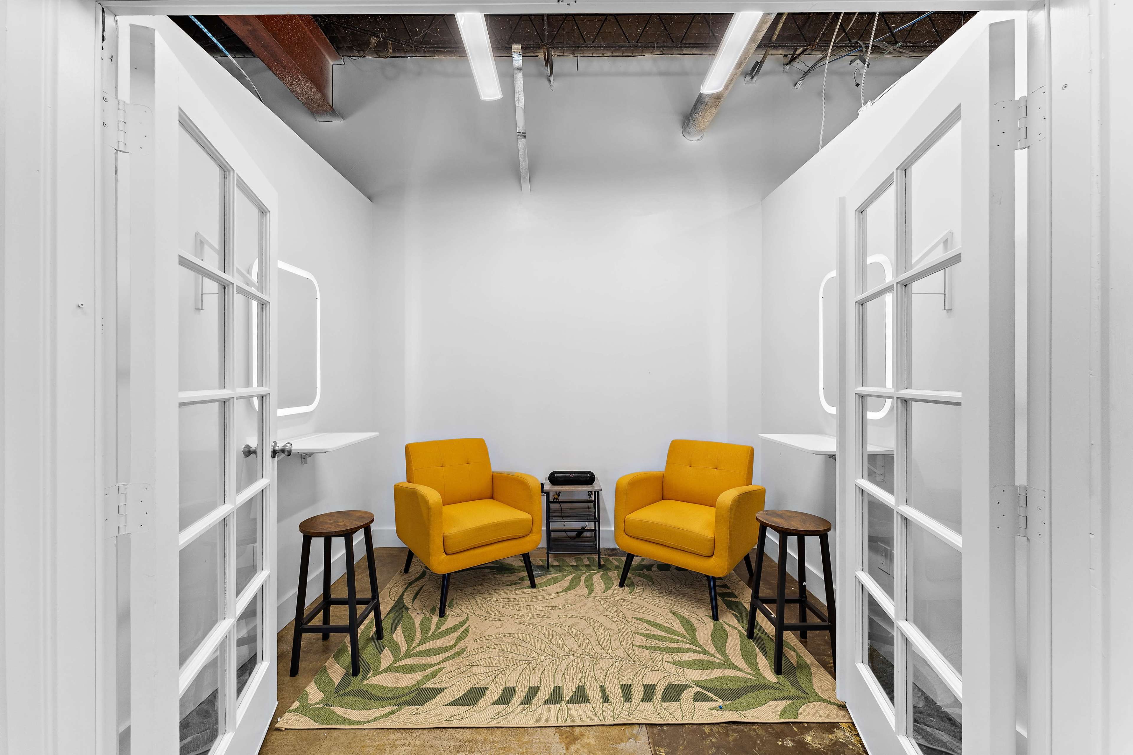 The image shows a small room divided into two sections, each featuring a yellow armchair, a small black table, and a green leaf-patterned rug on the floor.